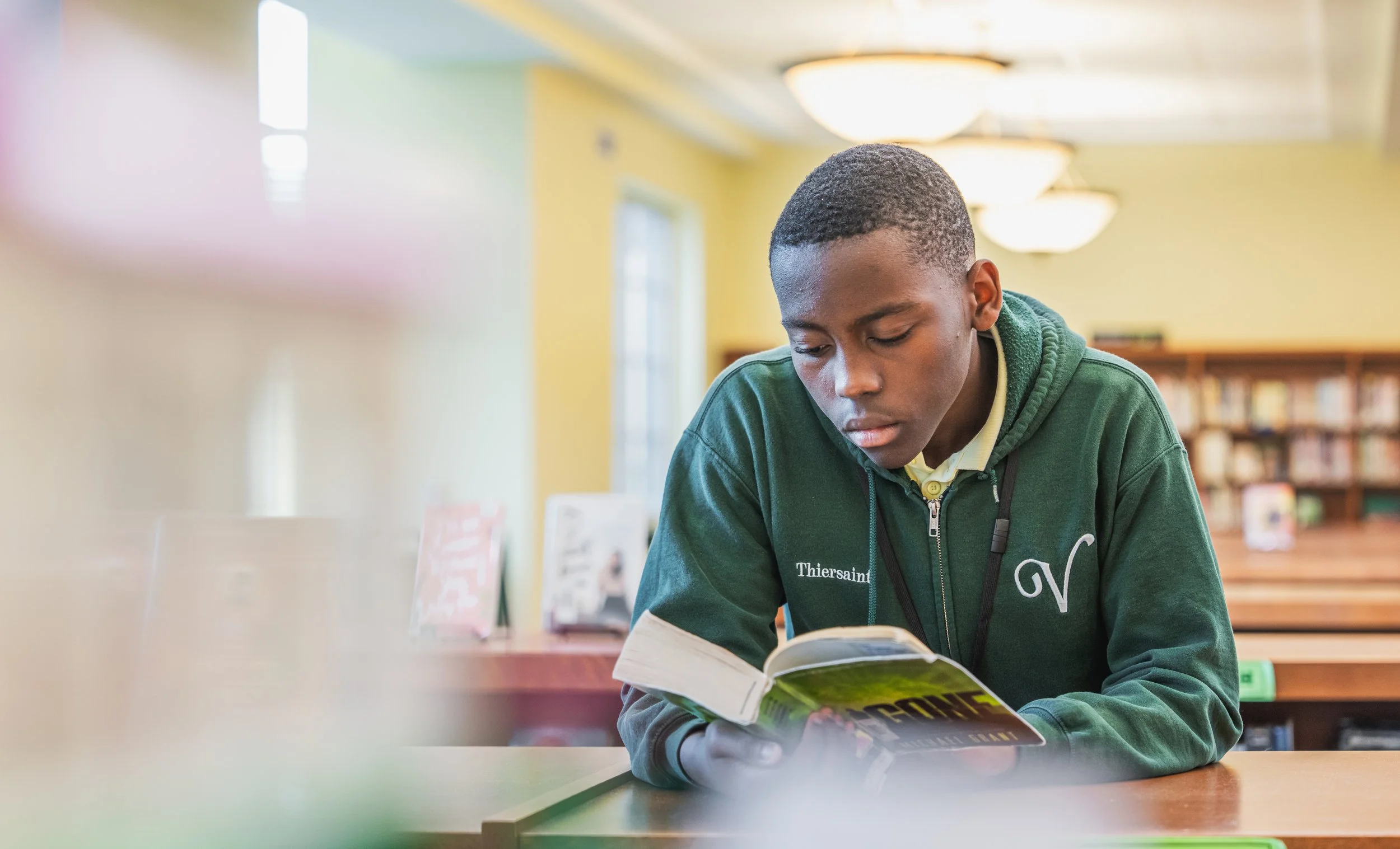 A young man in a green hoodie reading a book at a library.