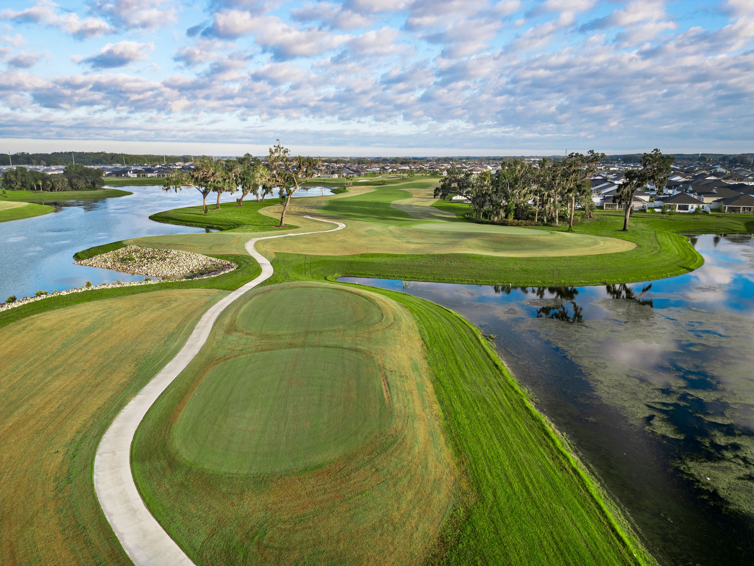Aerial view of a golf course with water hazards, green fairways, sand traps, trees, and a residential area in the background.
