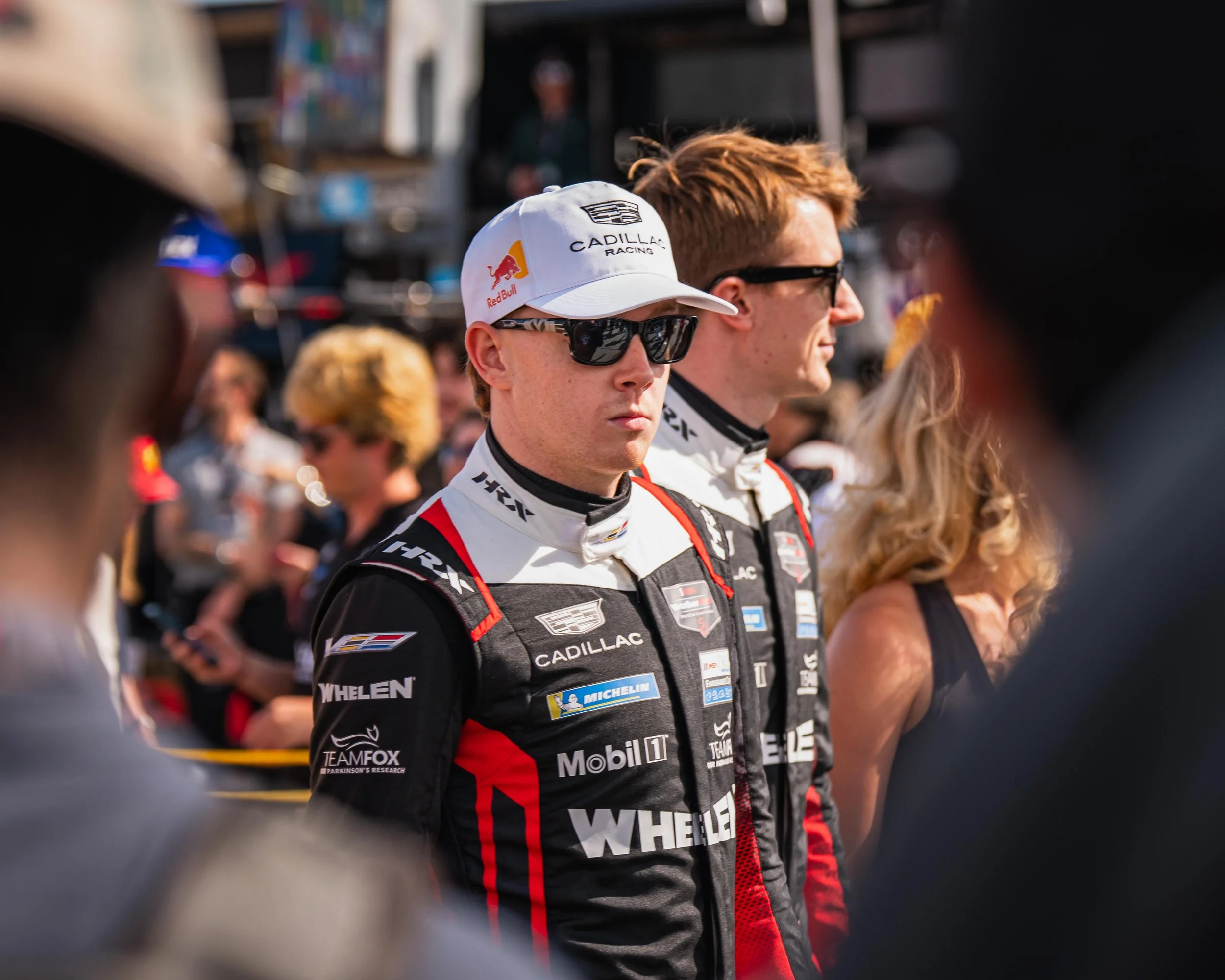 Race car drivers wearing black racing suits with sponsor logos, one with sunglasses and a white cap, standing in a crowded outdoor setting.