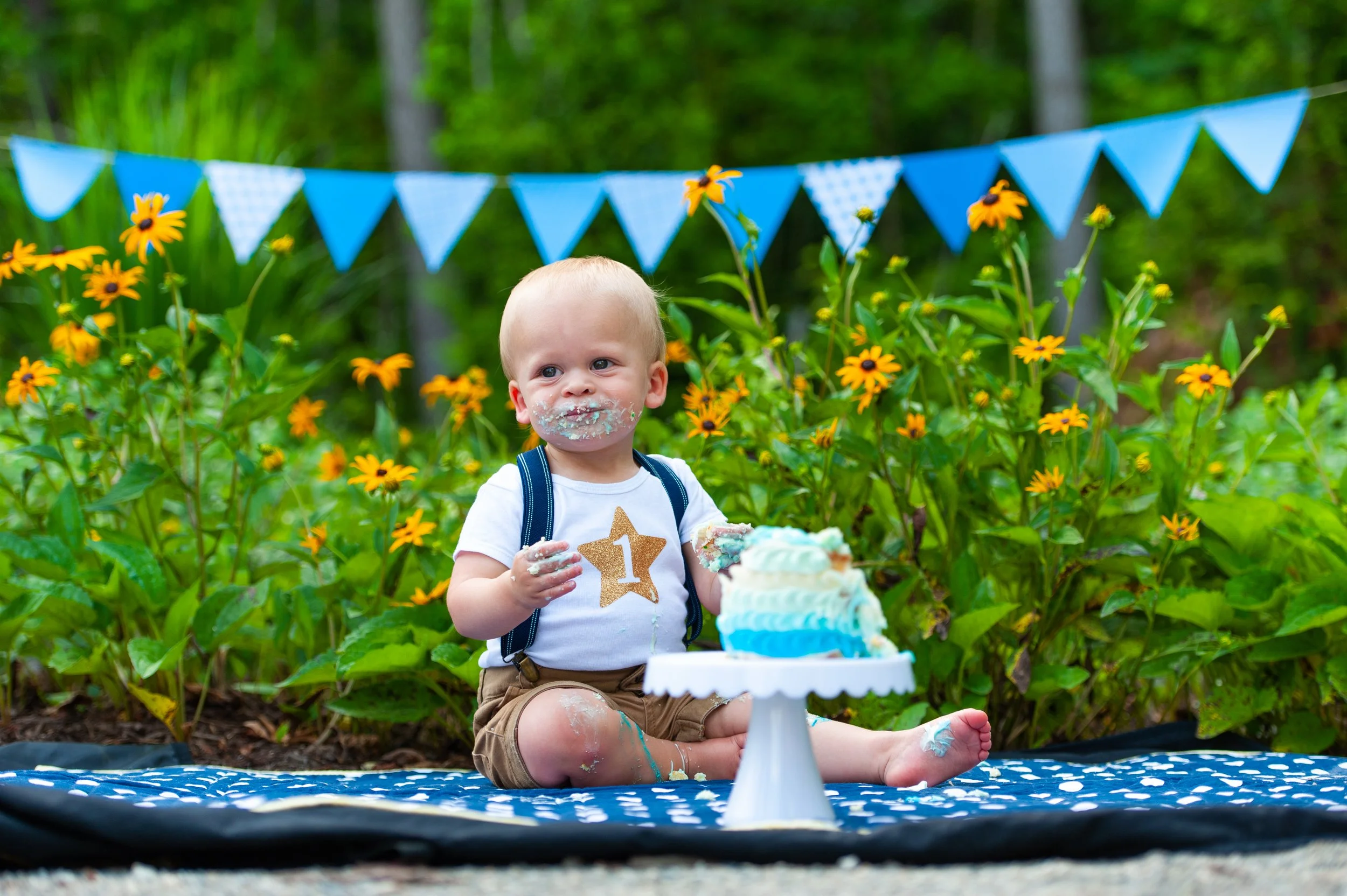 A young boy at his first birthday party sitting outdoors on a blanket with a partially eaten cake in front of him. He has cake on his face and hands, wearing a white shirt with a gold glitter number one, and a blue backpack. There are yellow flowers 