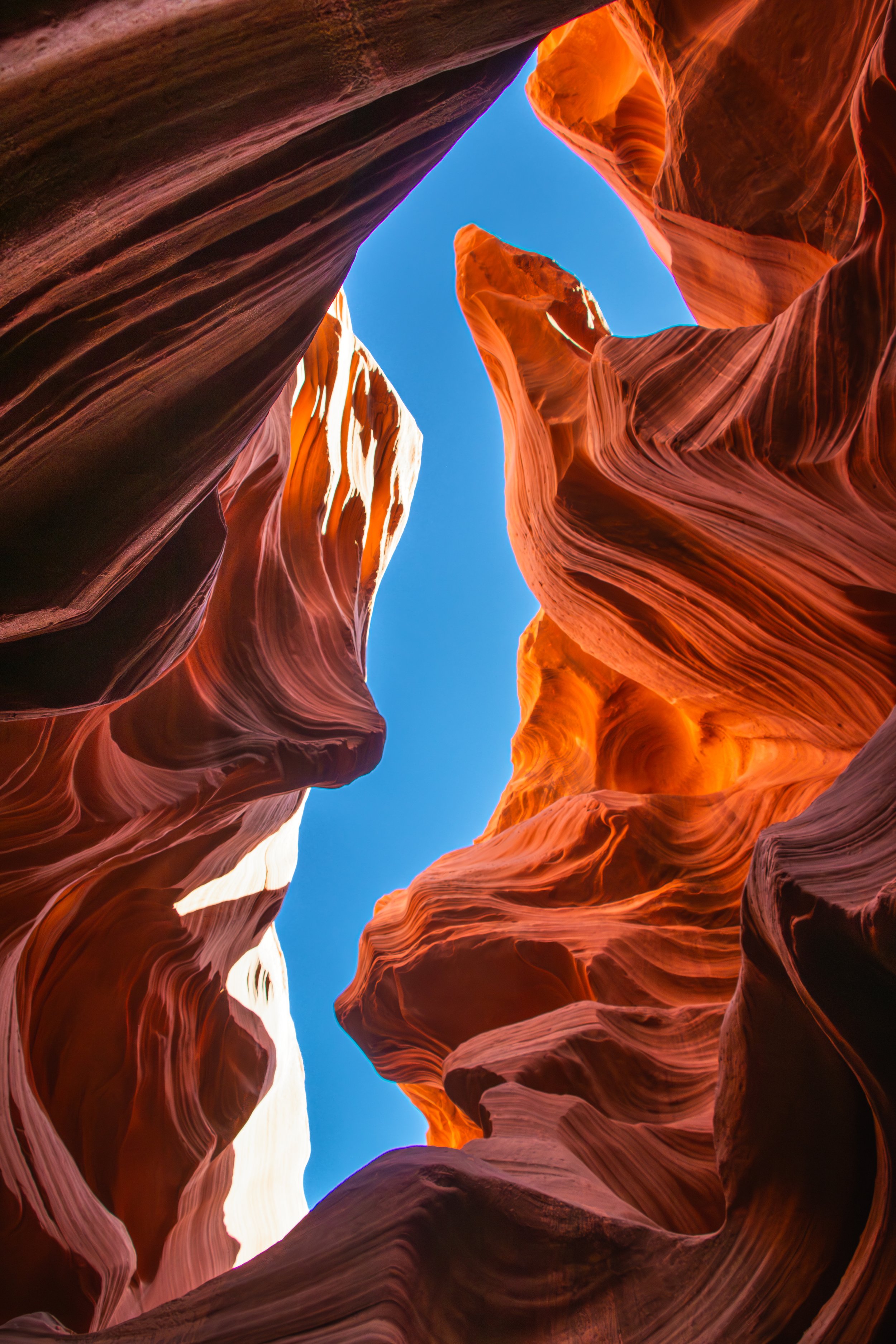 View from the bottom of antelope slot canyon with colorful, smooth, layered rock formations and a clear blue sky overhead. 