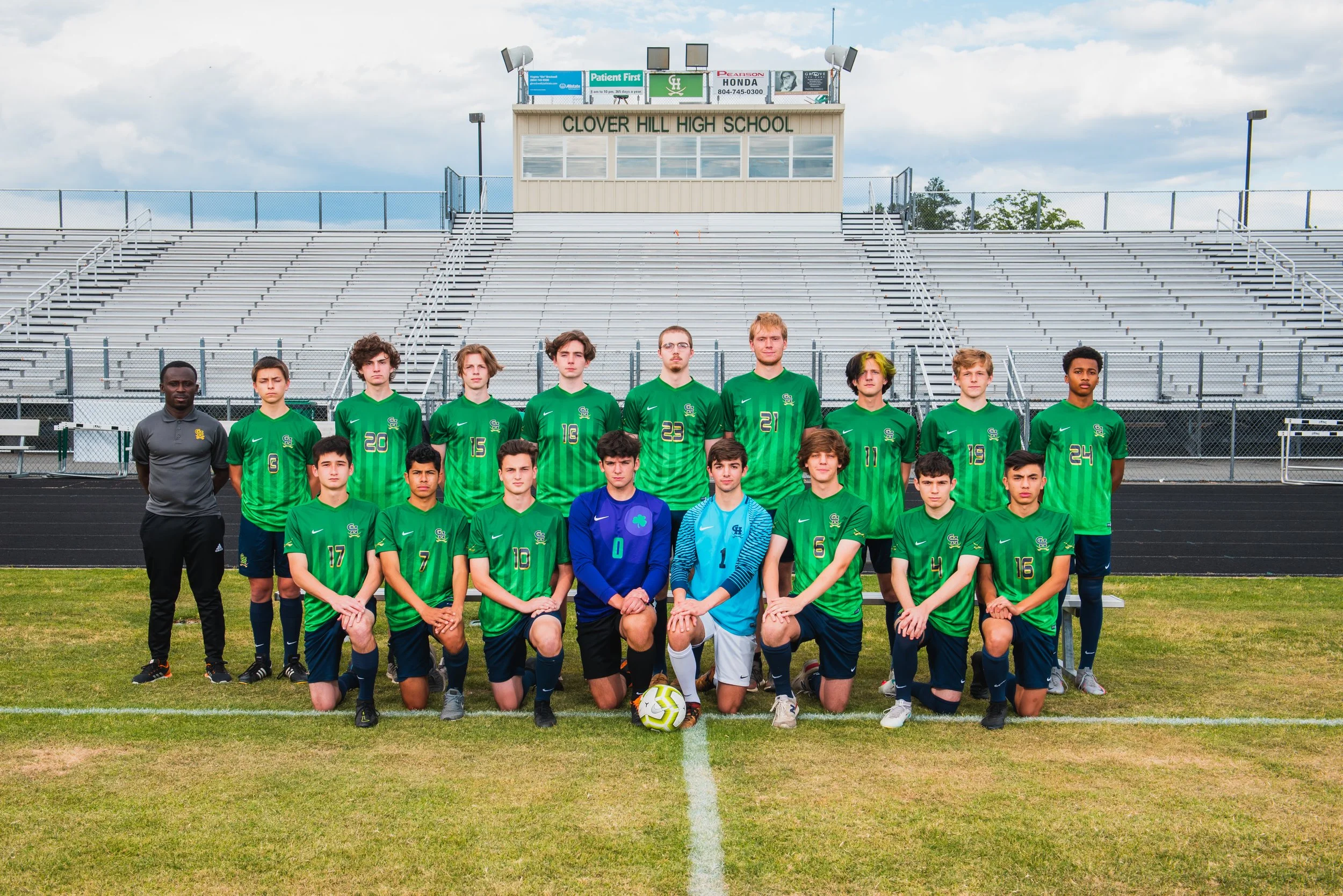 A soccer team of young male players and their coach, dressed in green jerseys, posing for a team photo on a sports field in front of empty bleachers at Clover Hill High School.