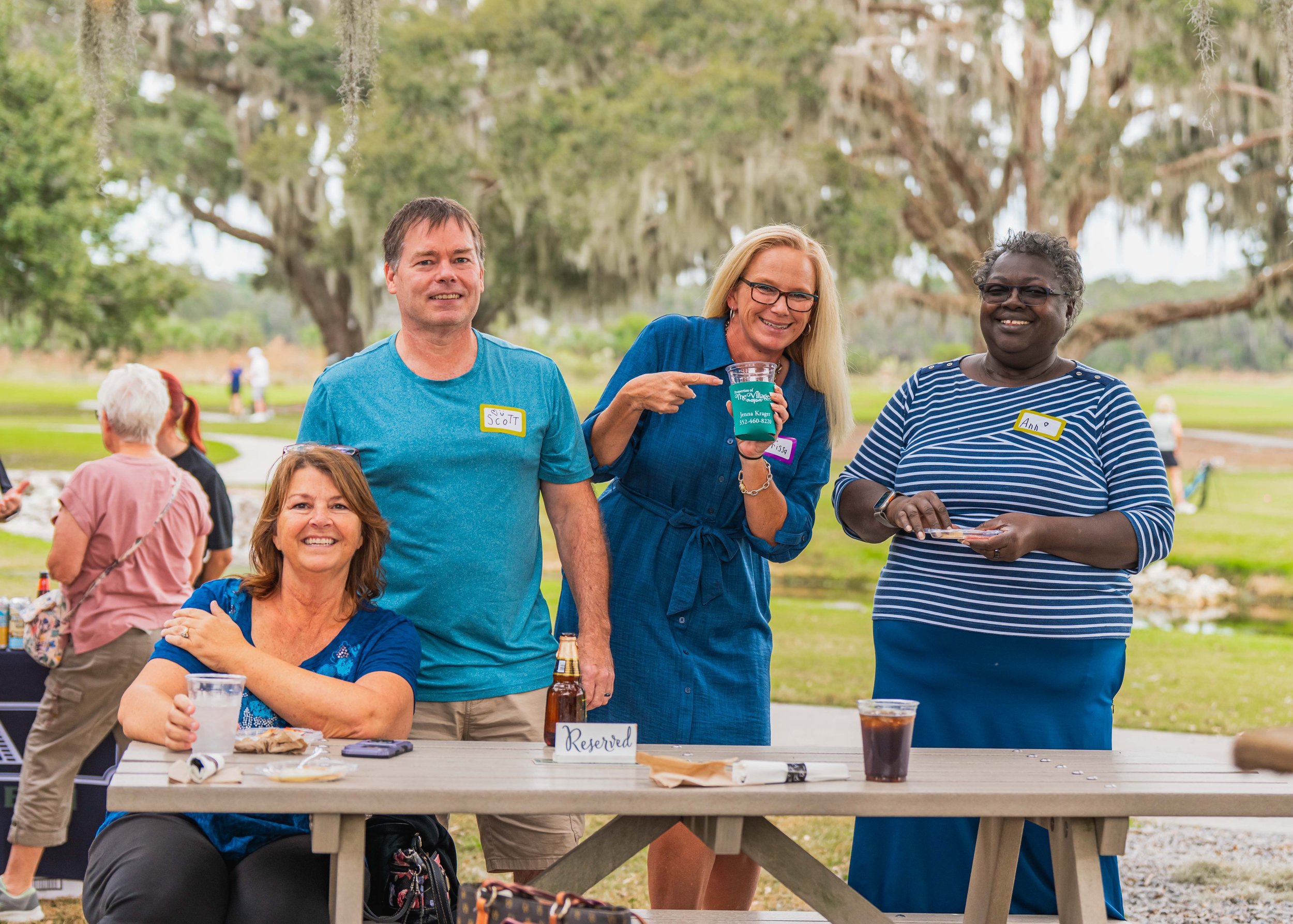Four adults standing outdoors, smiling, with a picnic table in front of them, with a woman holding a mug and a woman with a plate, in a park with large trees and a pond in the background.