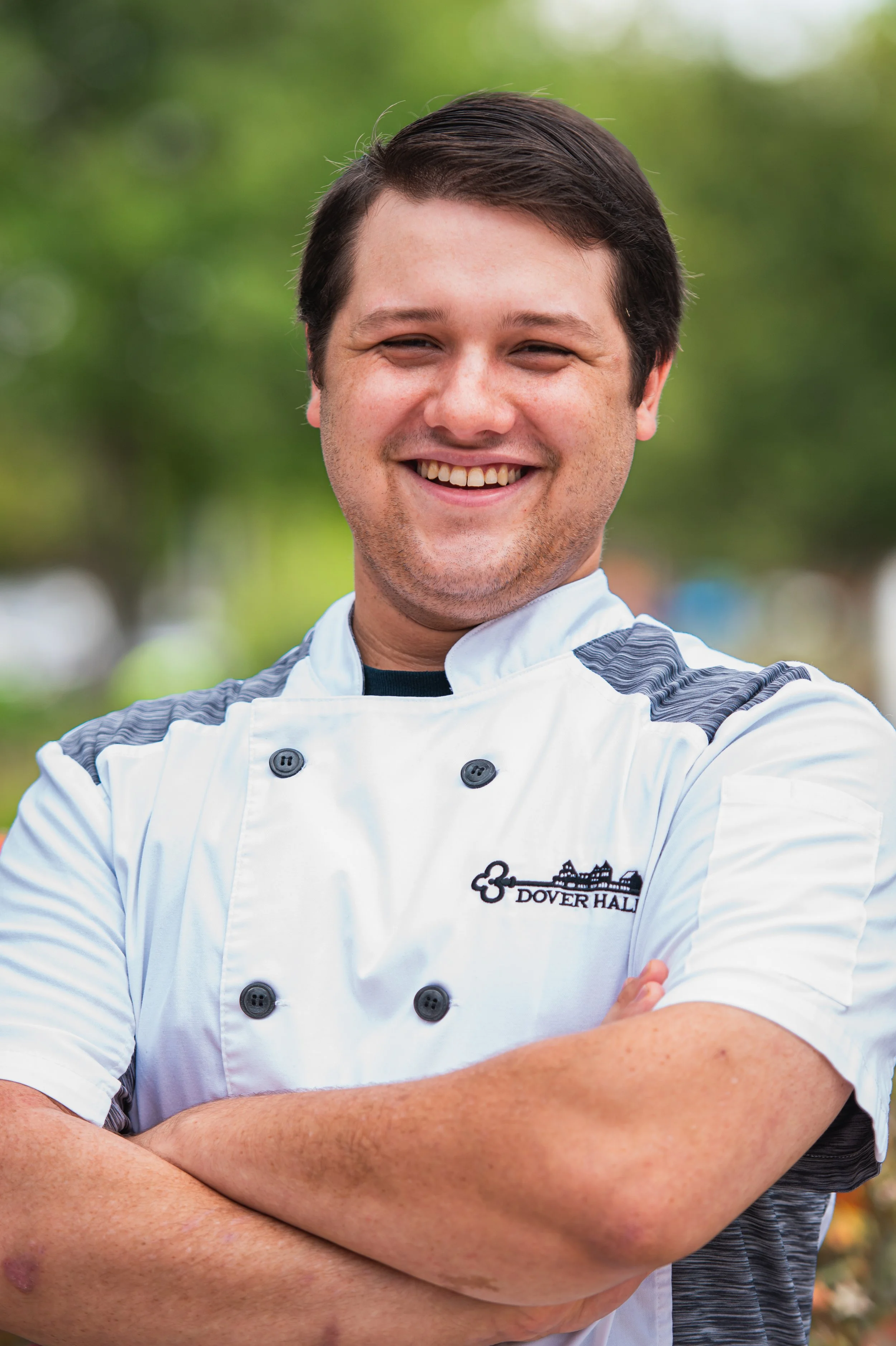 Portrait photography, Central Florida. Corporate headshots. A smiling man in a chef uniform with crossed arms, outdoors with blurred green trees in the background.