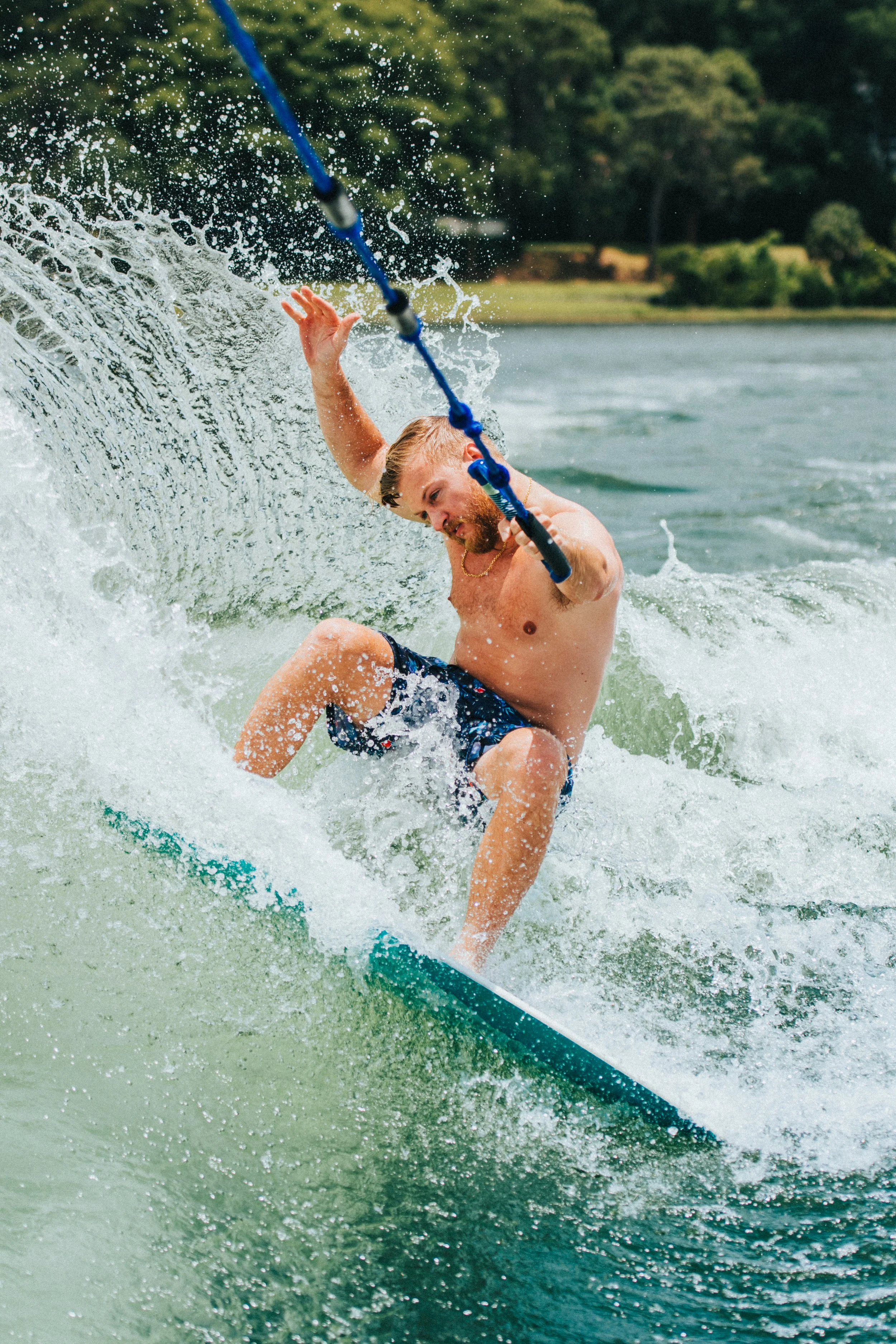 A man with a beard and long hair, wearing swim shorts, is wakeboarding on a river, holding onto a tow rope as he leans back and balances on the board, with water splashing around him and a tree-lined riverbank in the background.