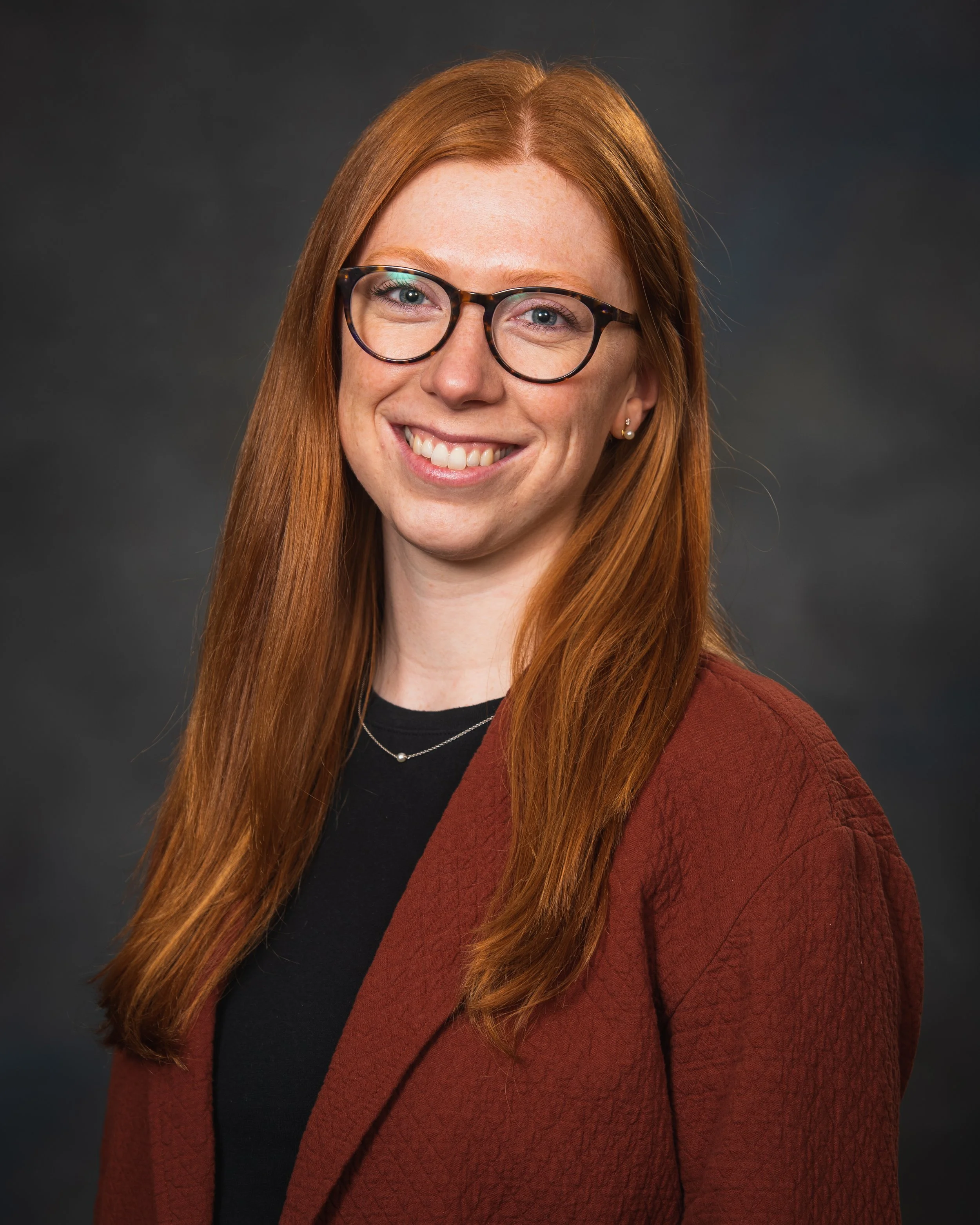 Portrait photography, Central Florida. Corporate headshots. A woman with long red hair, glasses, and a friendly smile, wearing a black top and a brown blazer, against a dark background.