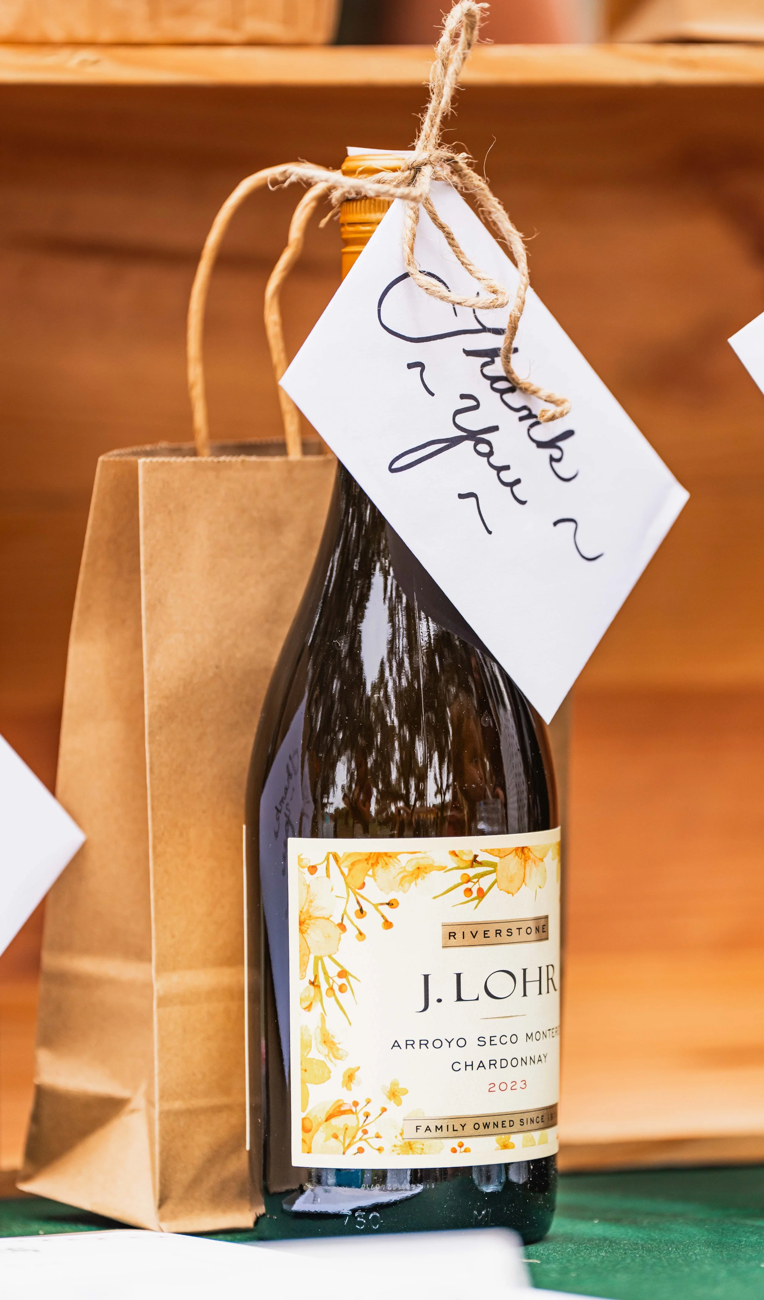 A bottle of J. Lohr Chardonnay wine with a gift tag that says "Thanks You" hanging from a twine string, placed beside a brown paper bag.