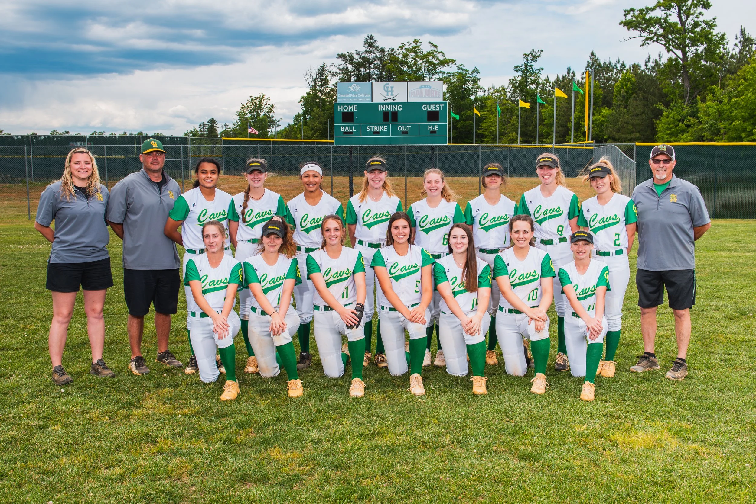 A girls' softball team with coaches posing on a baseball field for a team photo. The players are wearing white and green uniforms with the word 'Cavs' written on them, and everyone is smiling.