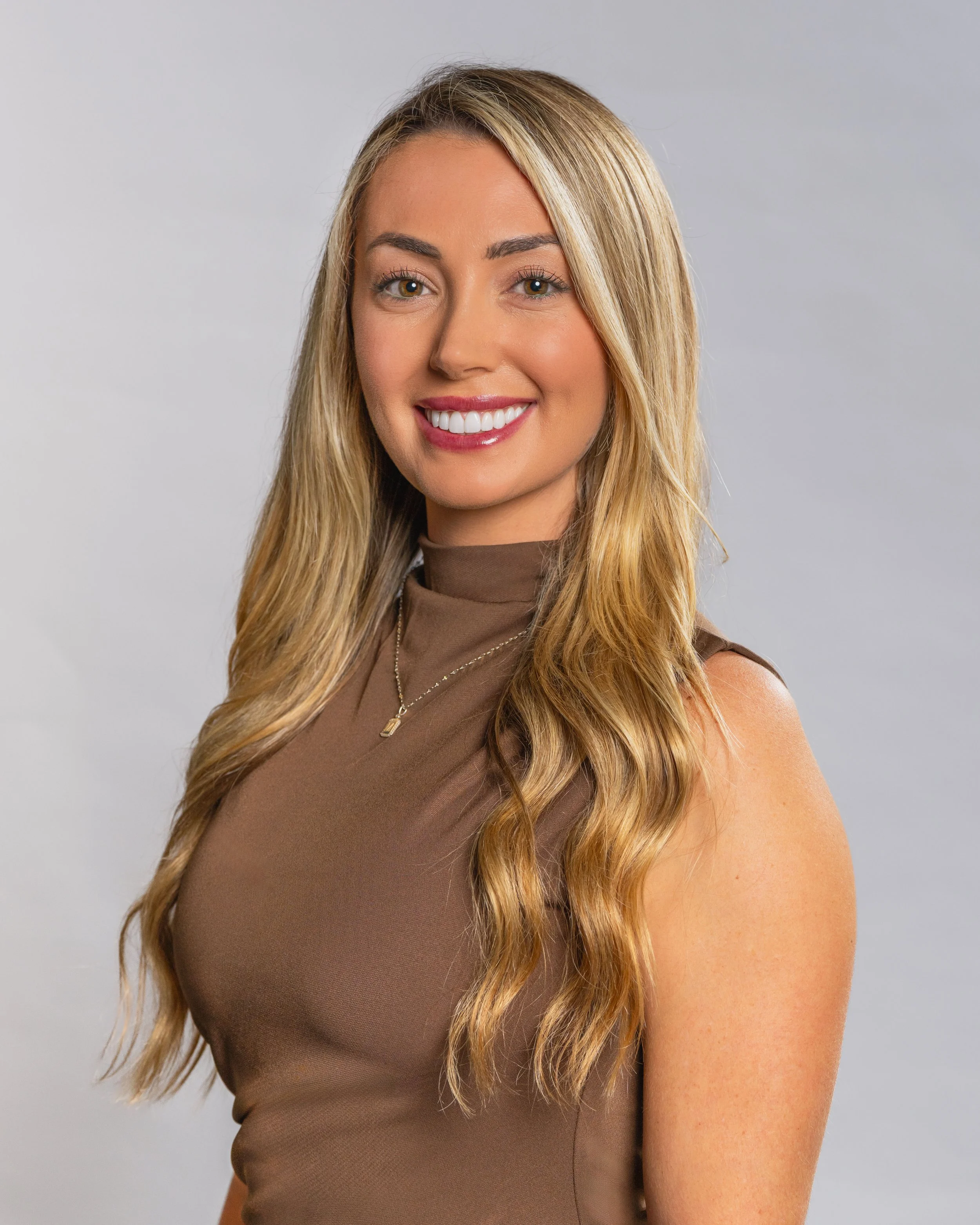 Portrait photography, Central Florida. Corporate headshots. A woman with long wavy blonde hair, wearing a sleeveless brown top, smiling against a plain light gray background.