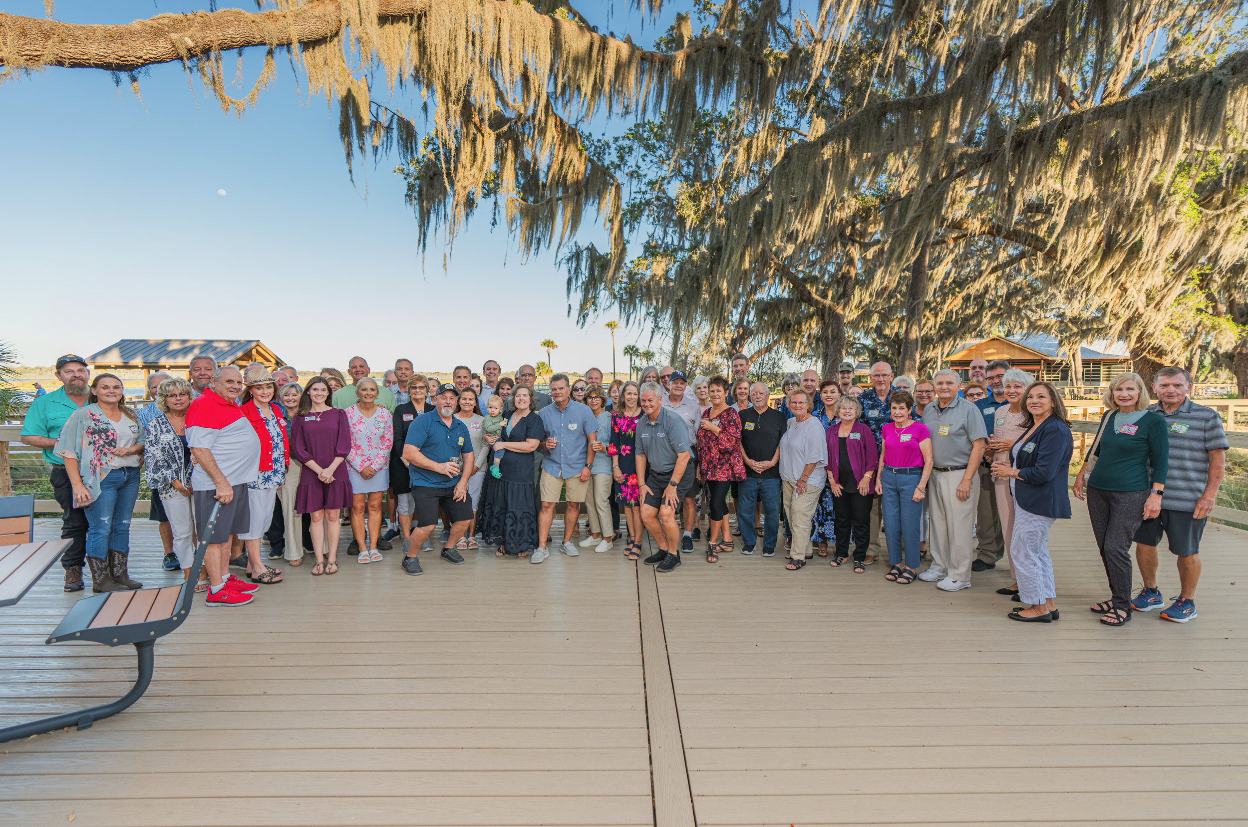 A large group of people gathered outdoors on a wooden deck under trees with hanging moss, posing for a group photo during daytime.