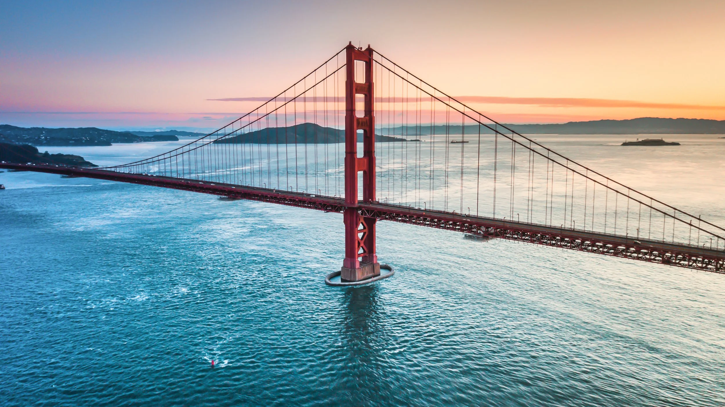 Aerial view of the Golden Gate Bridge at sunset with calm water below and distant landmasses in the background.