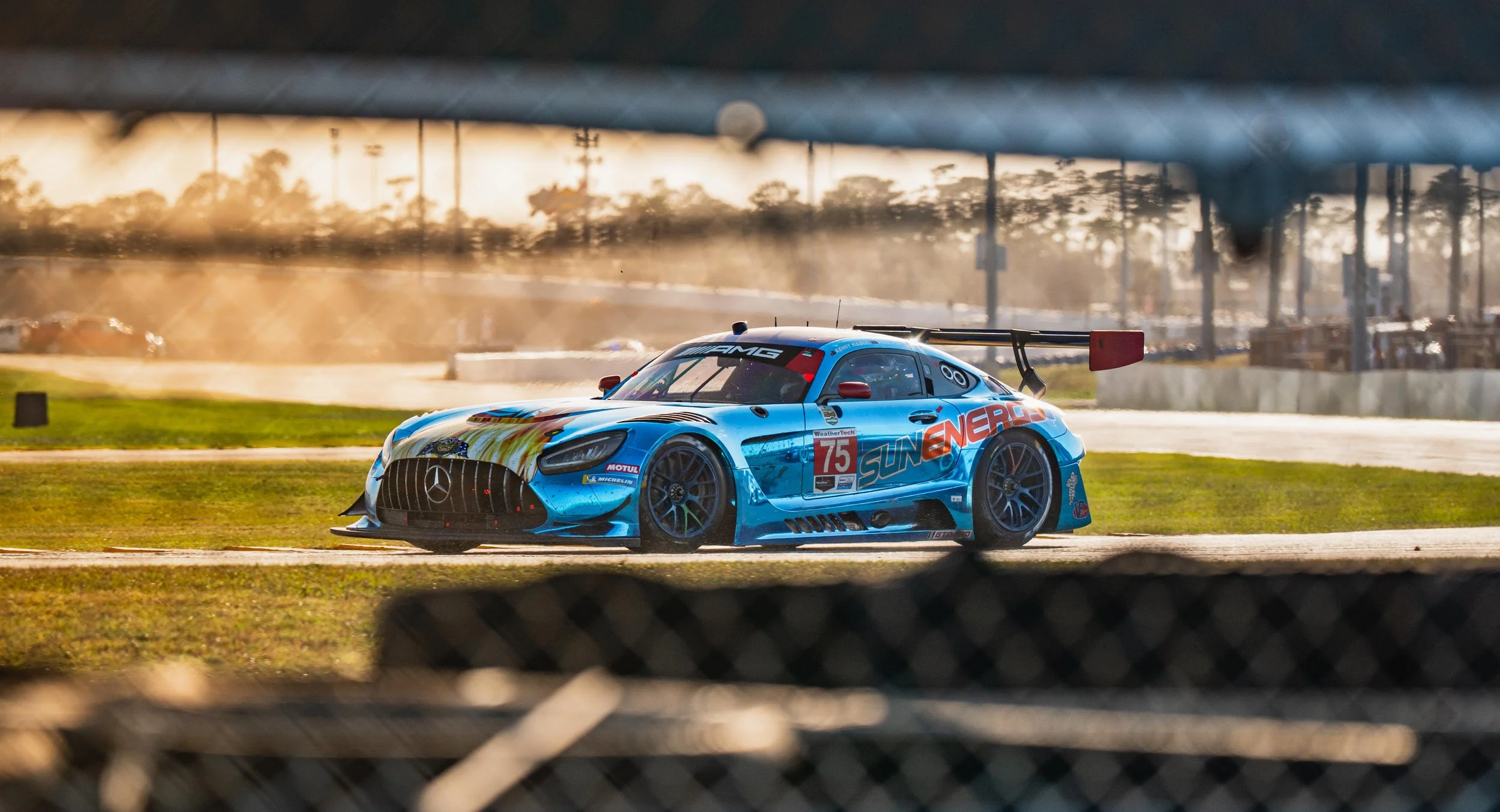 A blue race car on a track seen through a chain-link fence, with the sunset in the background.
