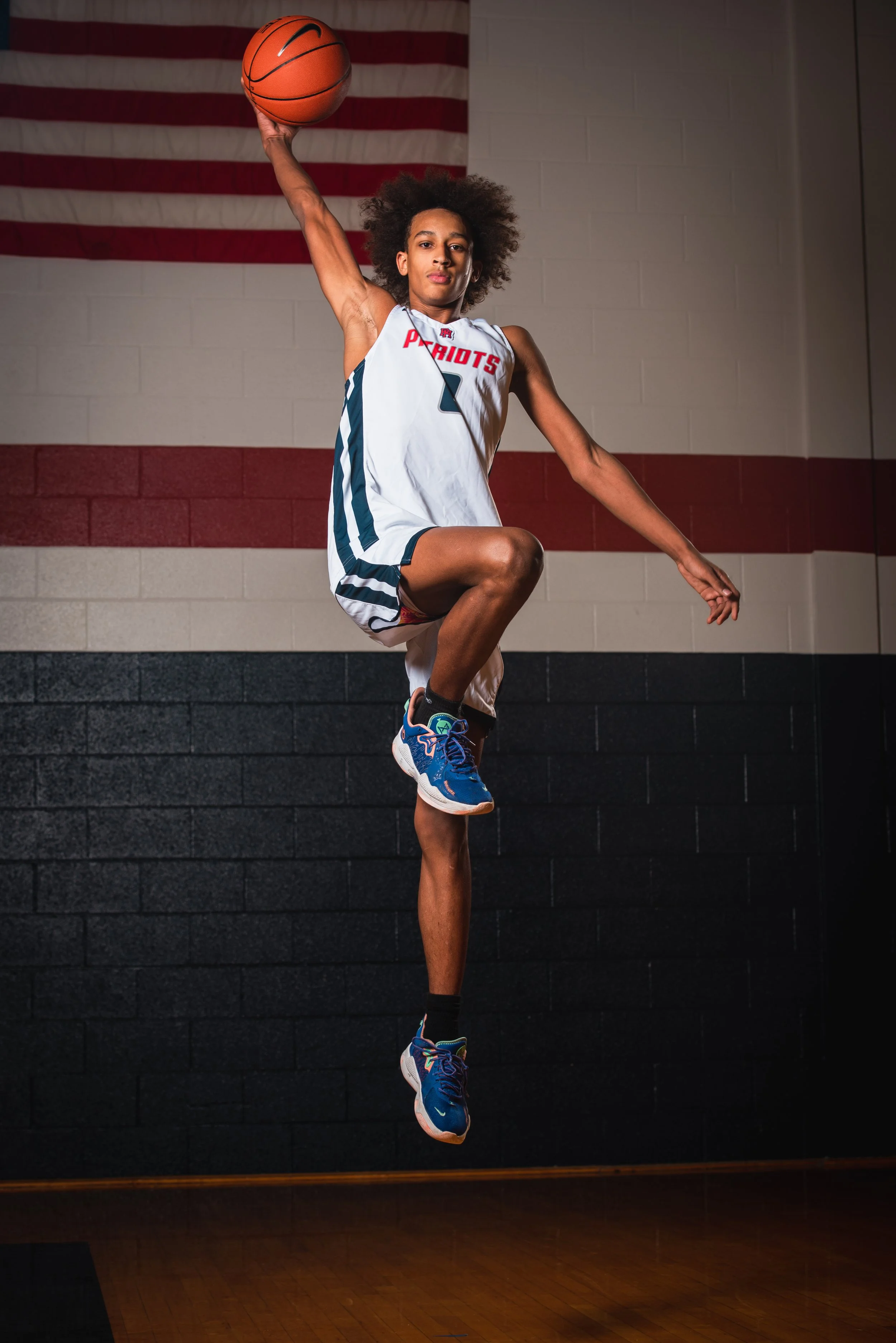 Male basketball player in a white jersey with 'Patriots' logo, jumping in gymnasium with an American flag on the wall behind him. School sports photography. Senior photos. 