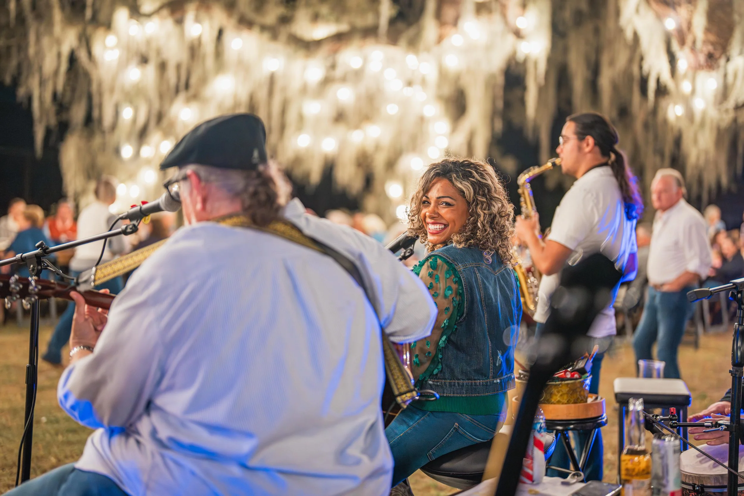 A woman with curly hair smiling while sitting with musicians at an outdoor event during the night, with hanging lights and trees in the background. Autumn Gold music