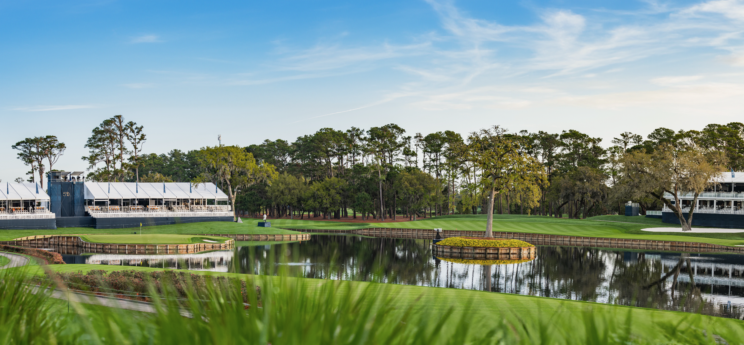 A scenic TPC Sawgrass golf course with green fairways, water hazards, and surrounding trees, with a clubhouse and outdoor seating area in the background. Island green on hole number 17. 