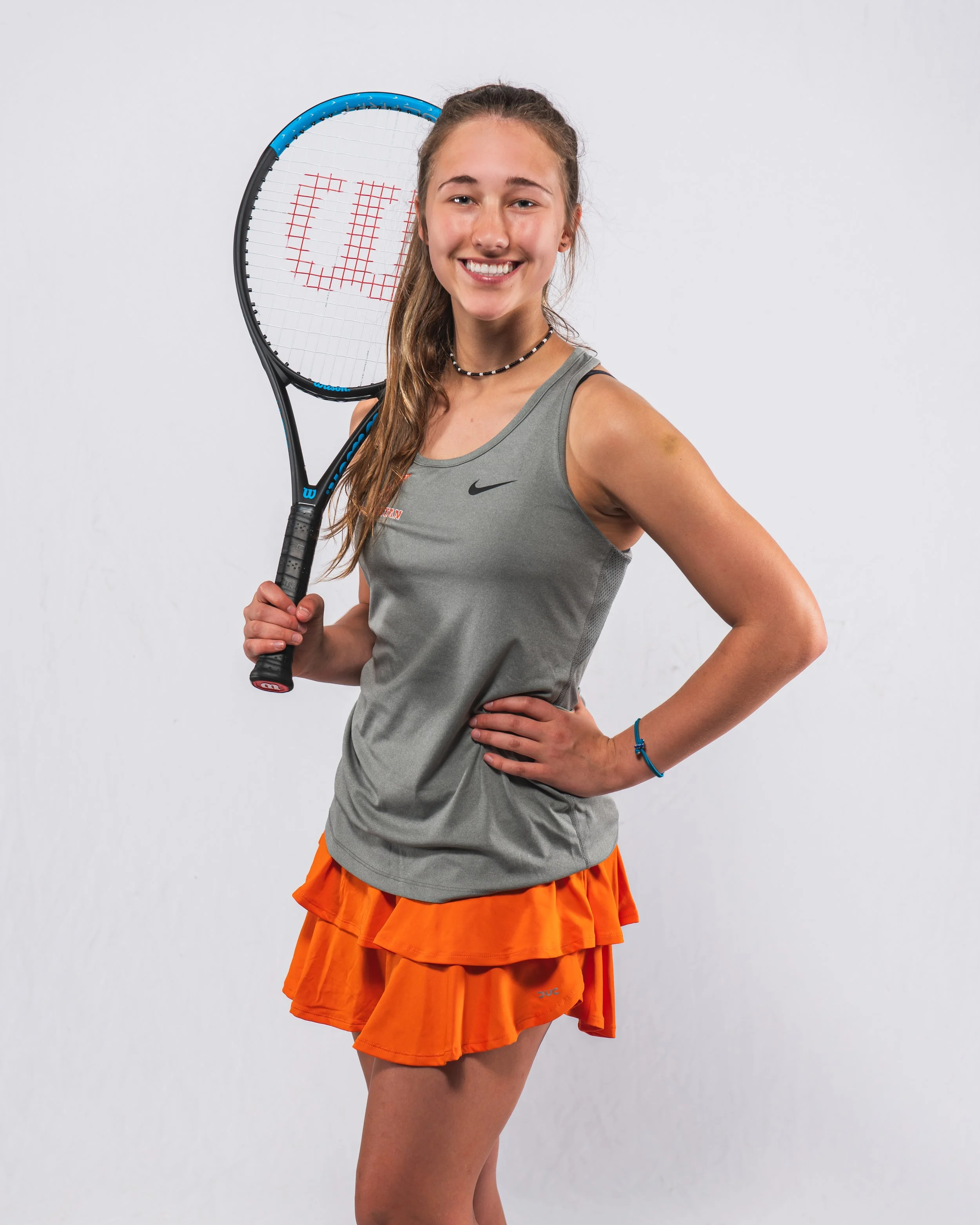 Young woman holding tennis racket with the letters 'USA' written on the strings, smiling, dressed in athletic wear, standing against a plain white background.