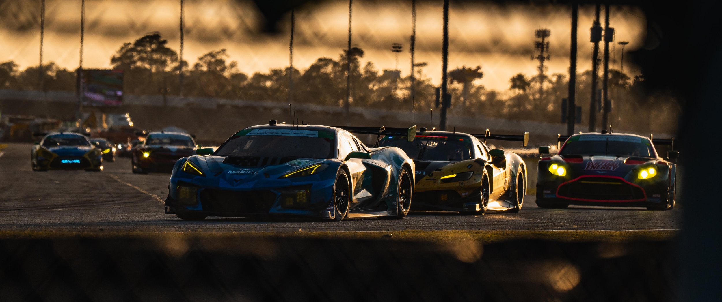 Race cars driving on a track during sunset with trees and power lines in the background.