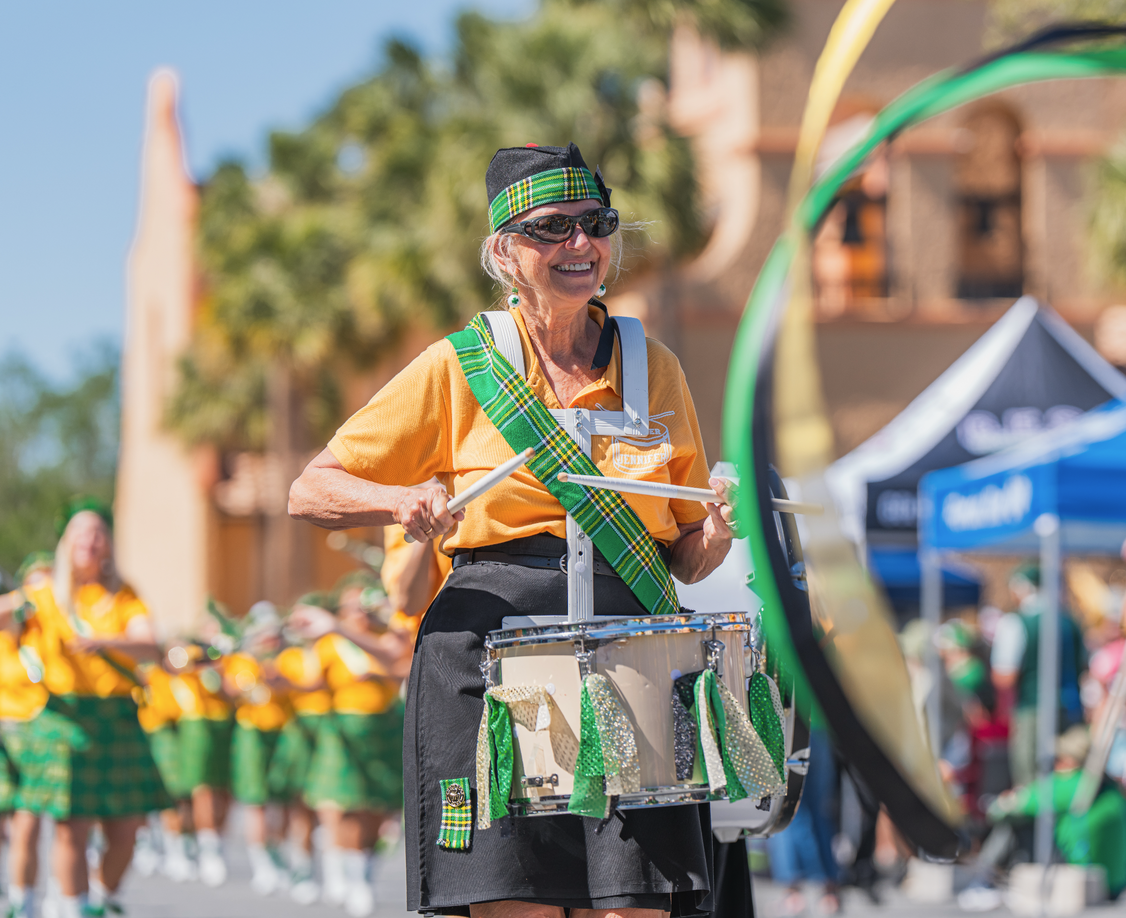 An elderly woman smiling while playing a percussion drum during a parade, wearing a black hat with a green tartan band, sunglasses, a yellow shirt with a tartan sash, and black skirt, with a group of dancers in similar attire in the background.