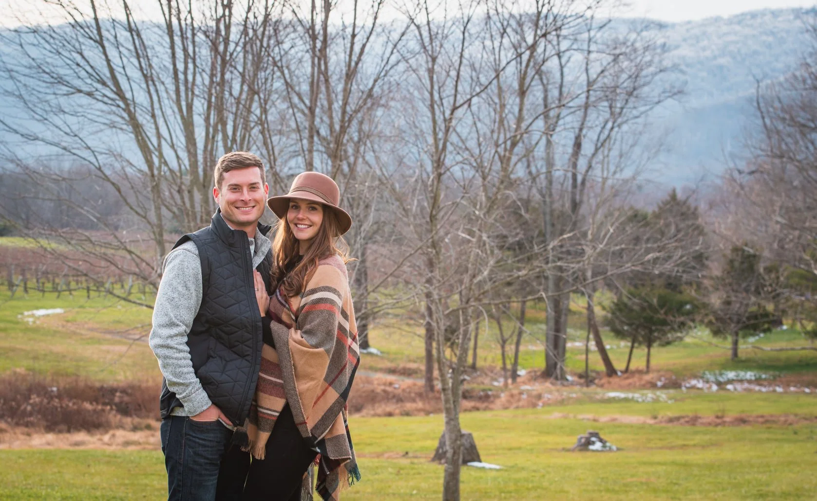 A smiling couple standing outdoors in a park-like area with trees and grass, dressed for cool weather.