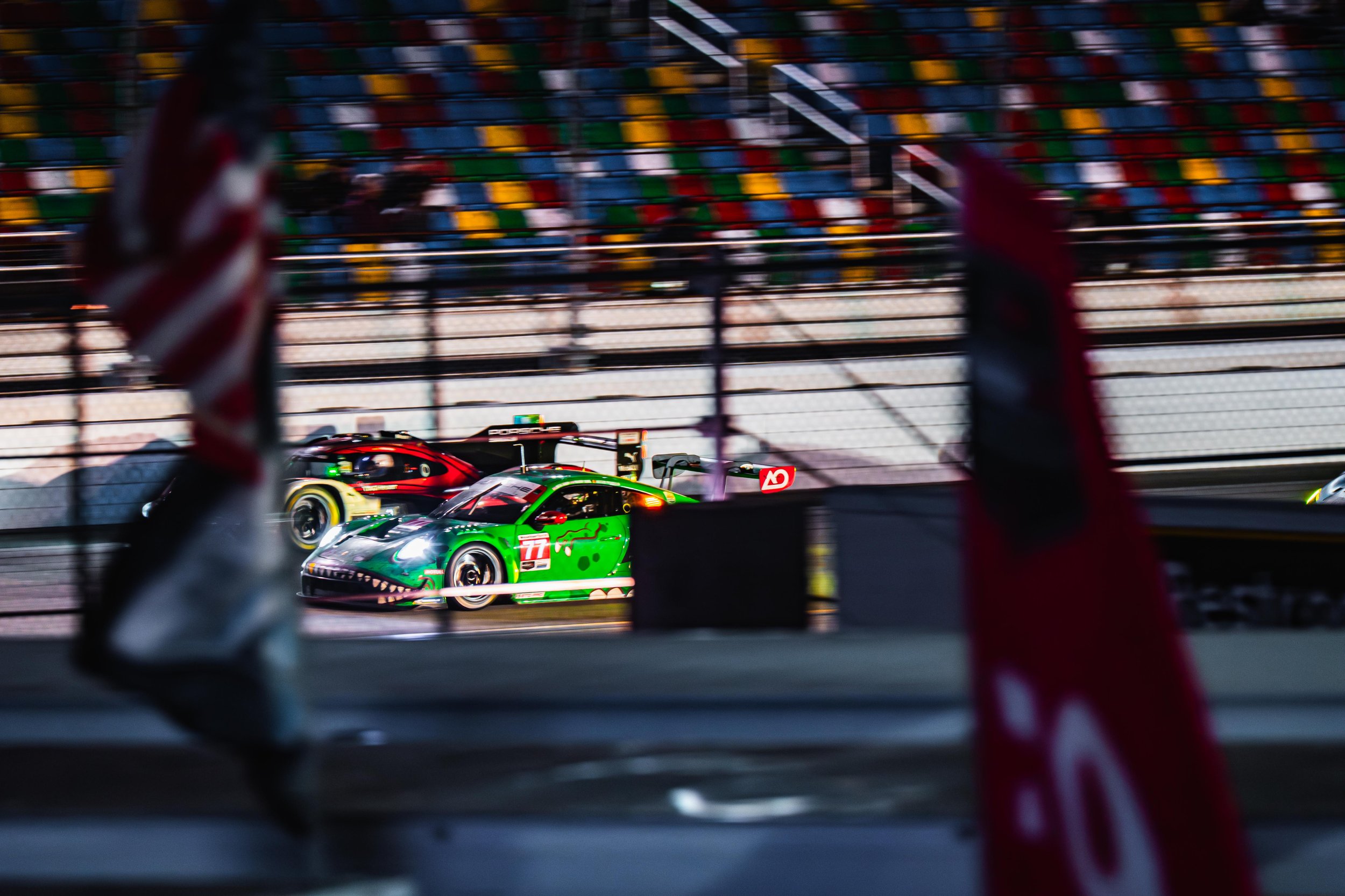 Two race cars on a track separated by a fence, with one car in the foreground and the other in the background, and colorful bleacher seats behind the track.