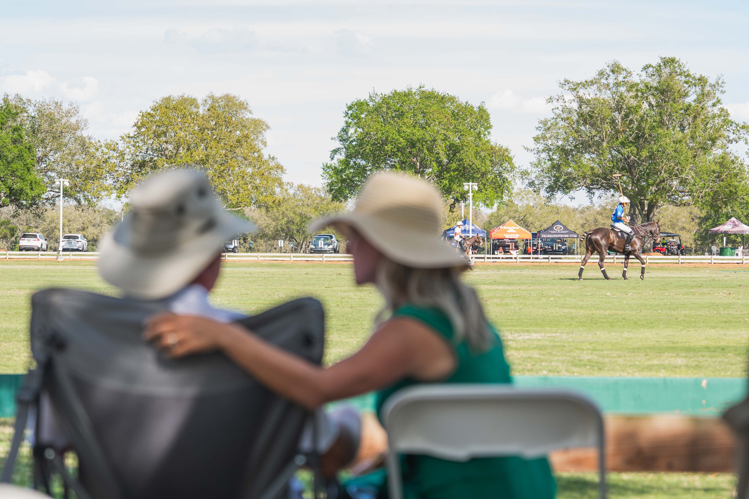 Couple sitting under a shady tree watching a polo match on a grassy field, with players on horseback and tents in the background on a sunny day.