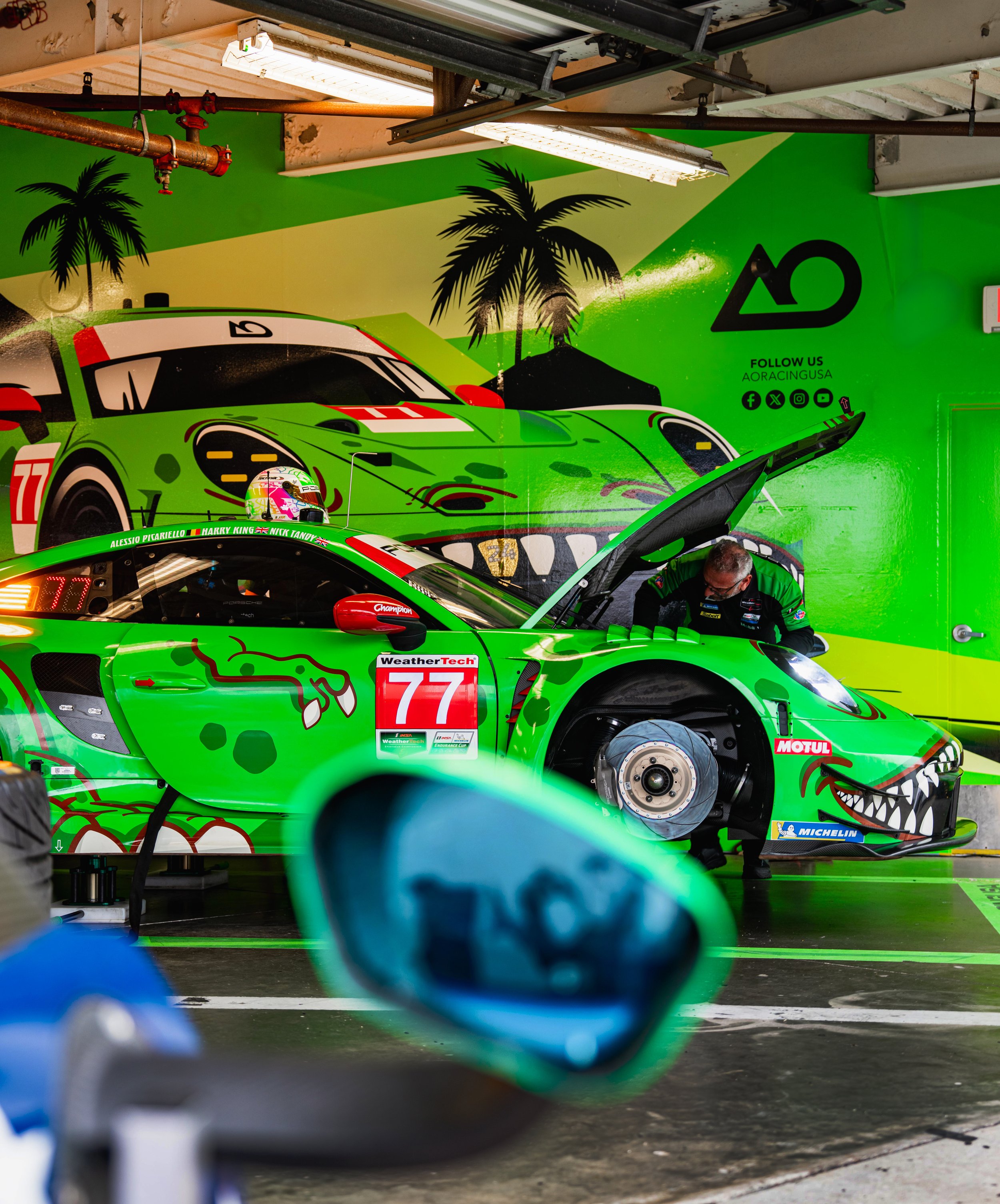 A race car with the number 77 on the side, painted in bright green with a dinosaur face design, is in a garage with a mechanic inspecting the engine. The garage walls are decorated with palm trees and a cartoonish alligator motif.