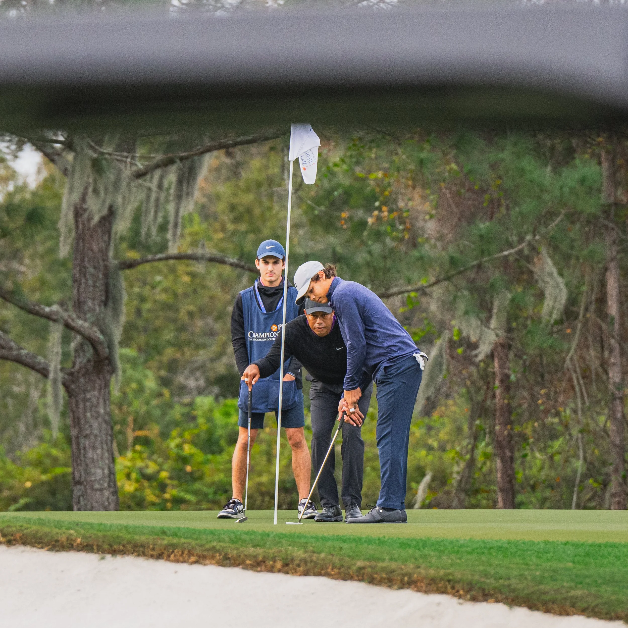 Tiger Woods and Charlie Woods practicing putting on a green surrounded by trees, with a caddy and another person assisting, seen from a view below the grass level.