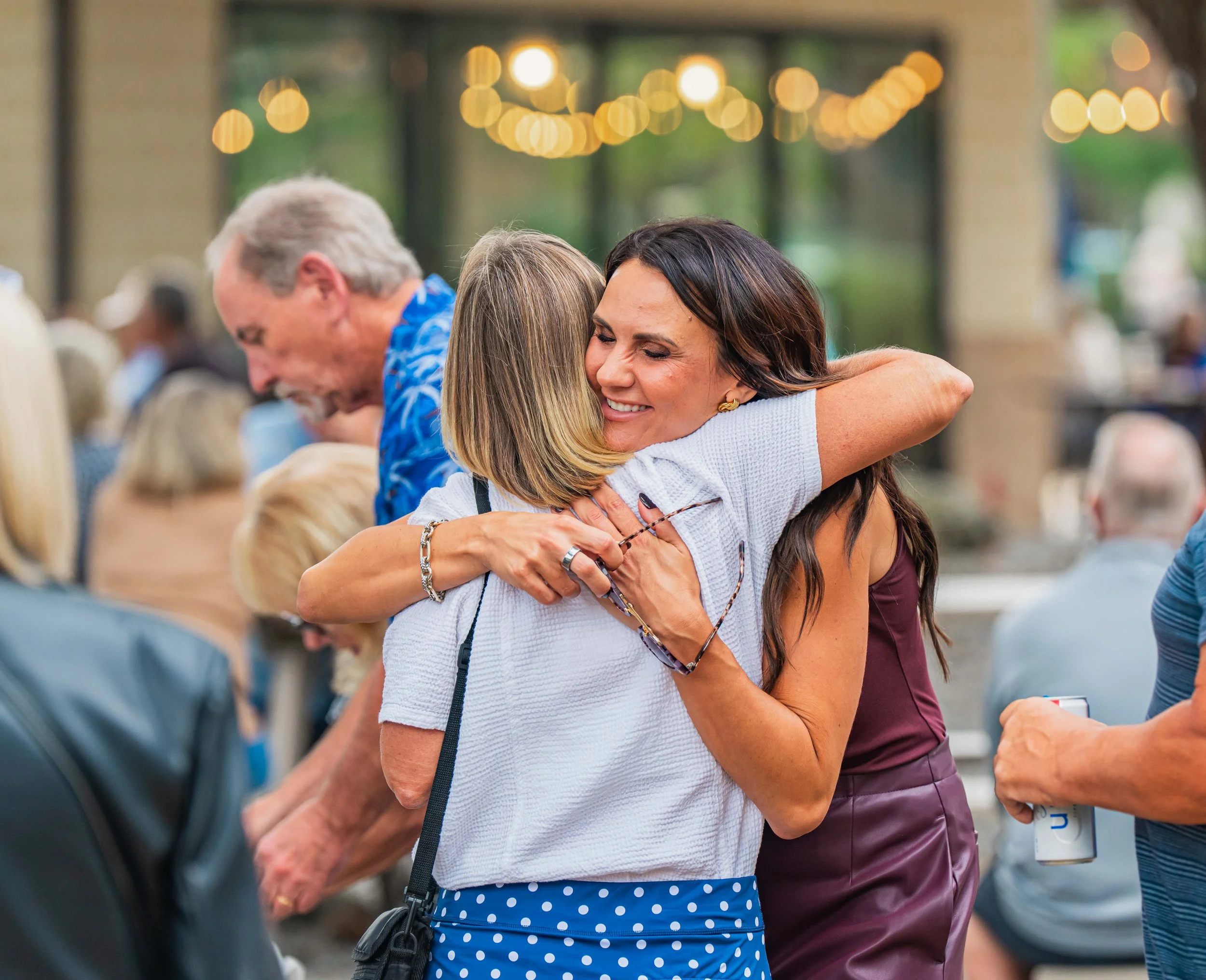 Two women hugging at a social gathering with other people in the background.