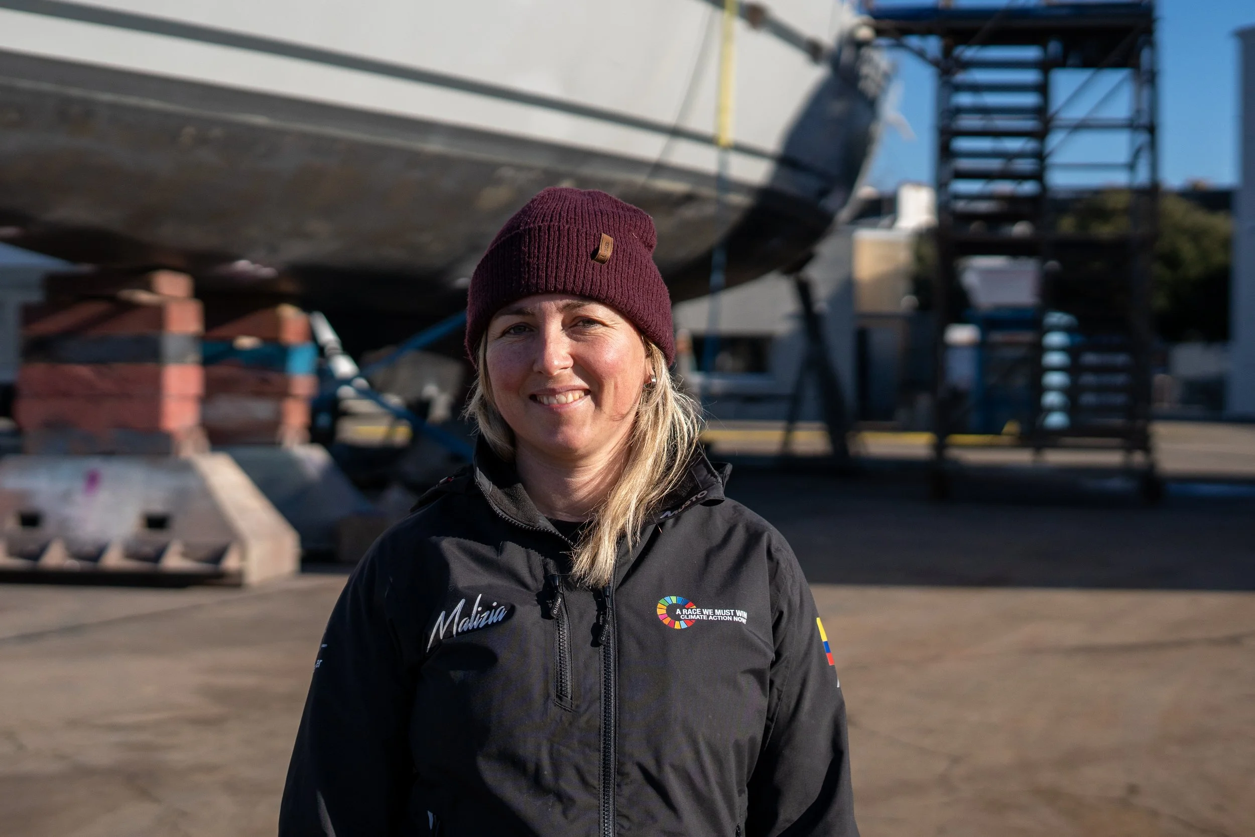 Oona Layolle and black jacket with sponsor patches stands outdoors near a large boat on a trailer, smiling at the camera.