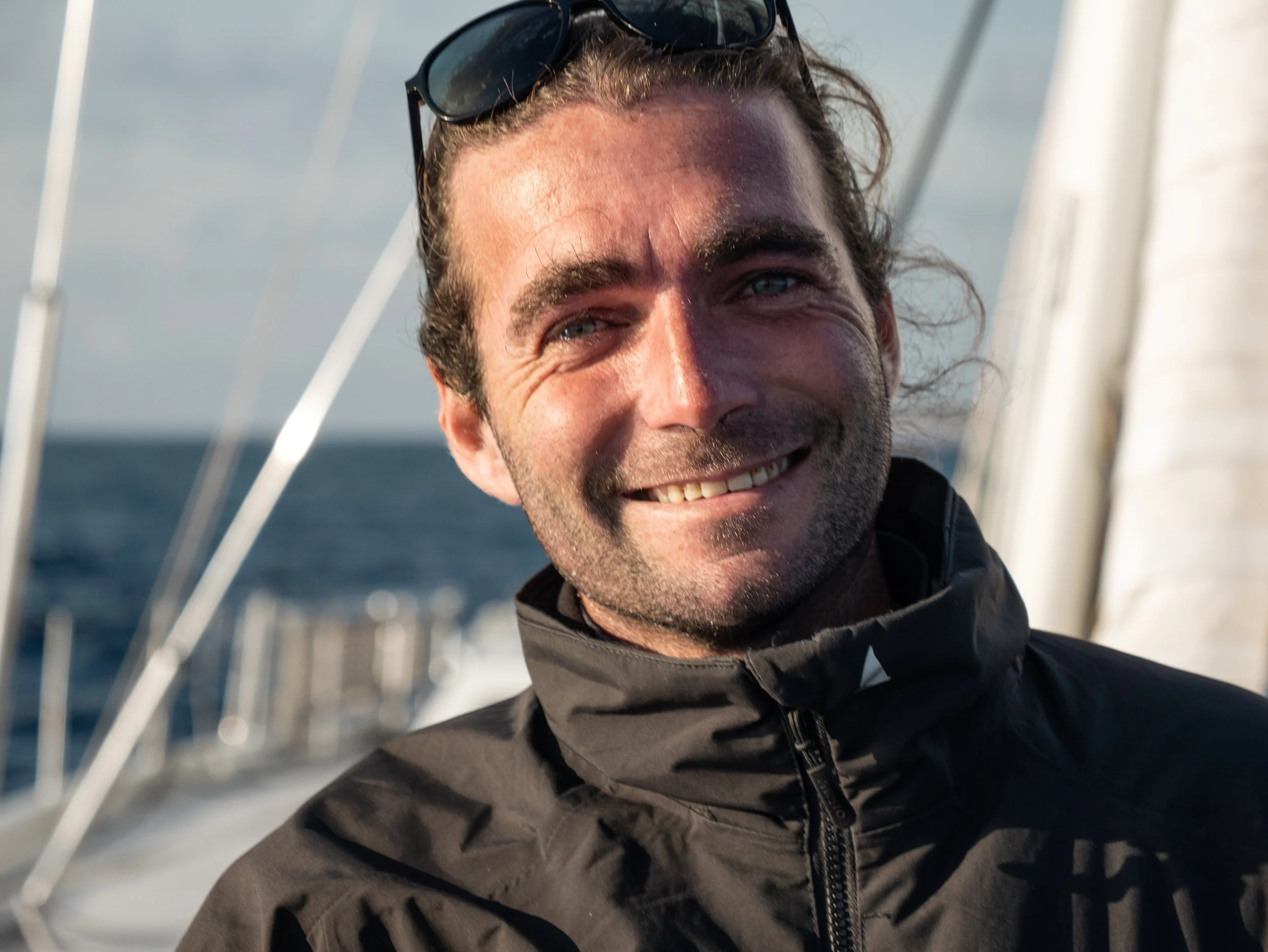 Smiling man wearing a black jacket and sunglasses on his head aboard a boat with the ocean in the background.