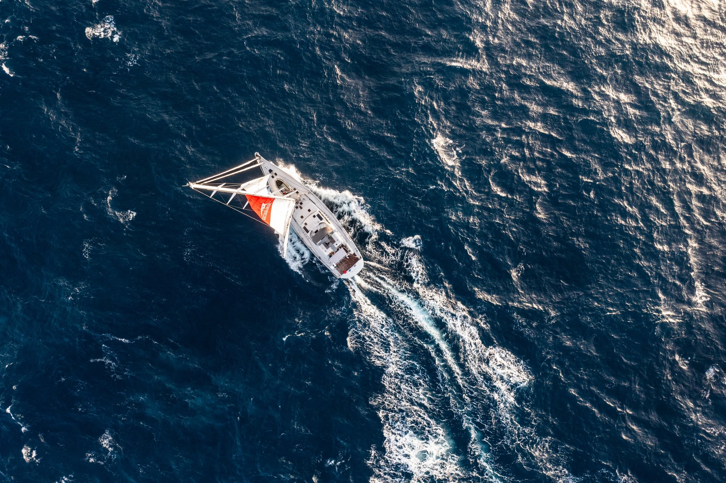 An aerial view of Malizia Explorer sailboat sailing on the open ocean, with visible waves and sunlight reflecting on the water.
