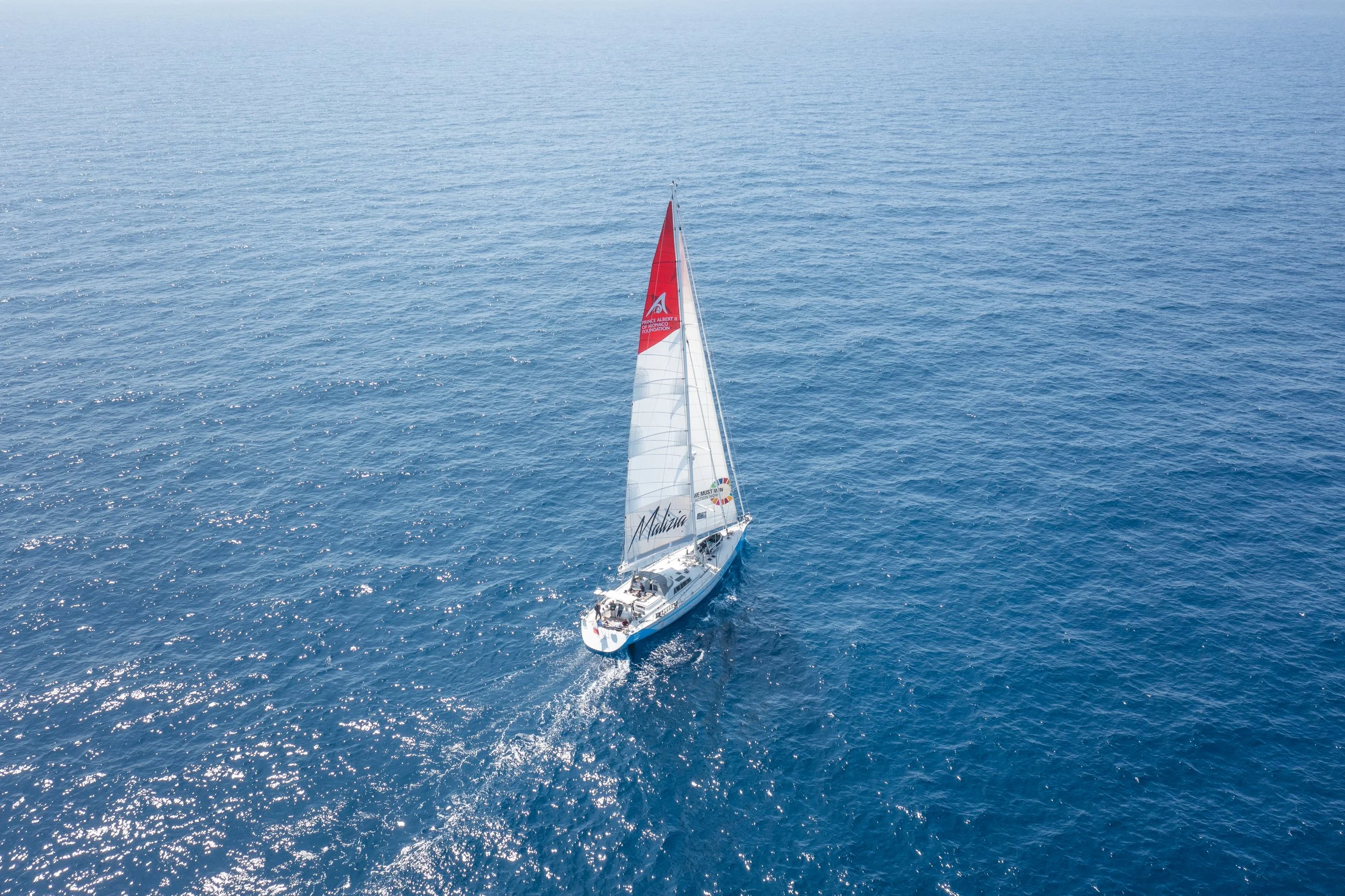 An aerial view of Malizia Explorer sailing yacht with white and red sails on calm blue ocean waters.