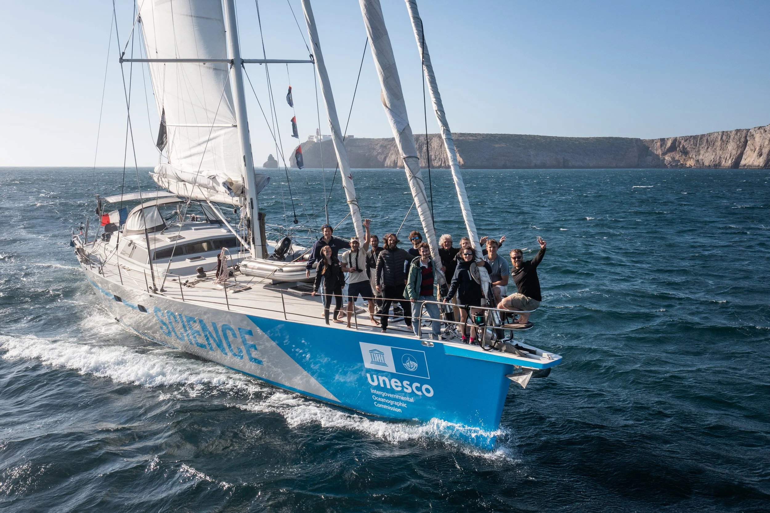 A group of people on a Malizia Explorer with visibility of the ocean and cliffs in the background, and the sailboat has UNESCO and other logos on its side.
