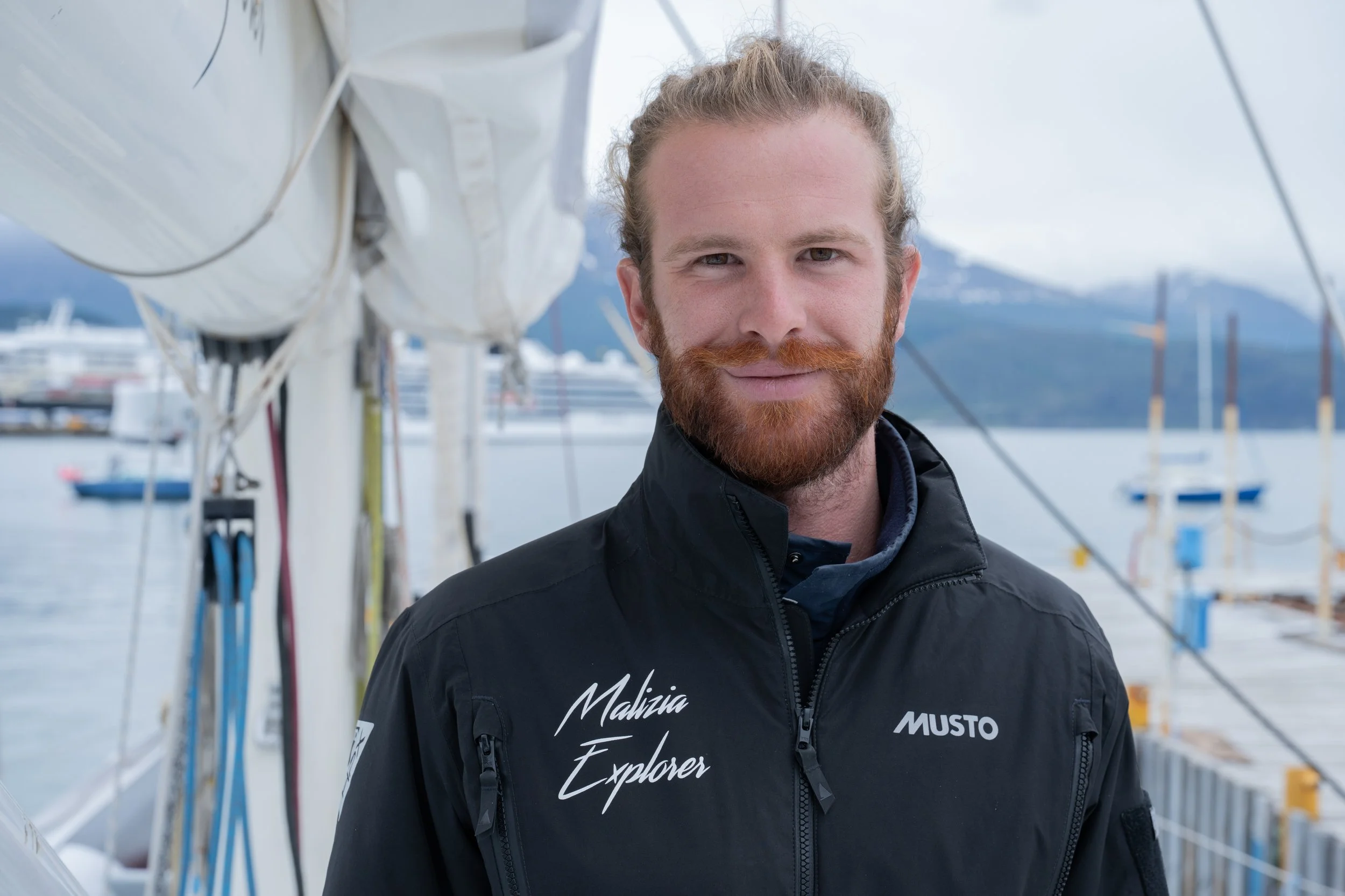 A man with a red beard and long hair tied back, wearing a black jacket with "Malizia Explorer" and "MUSTO" logos, standing on a dock with sailboats and mountains in the background.