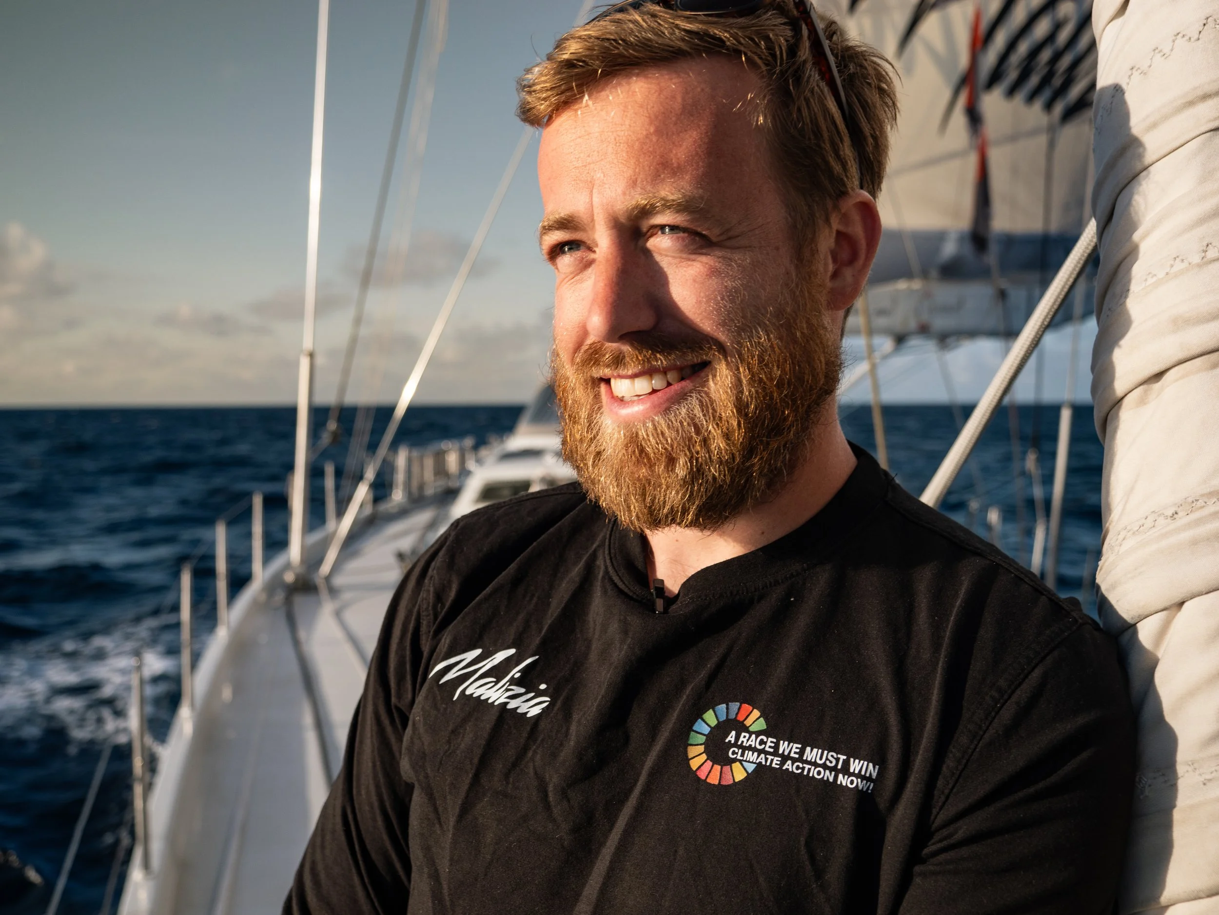 A man with a beard and short hair smiling on a sailboat with the ocean in the background during sunset, wearing a black shirt with a message about climate action.