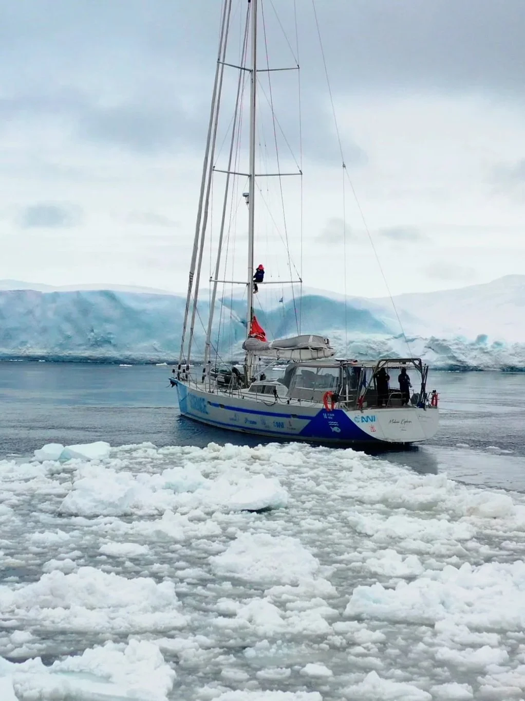 Malizia Explorer research vessel navigating icy waters with floating ice chunks, snow-covered mountains in the background, and an overcast sky, in Antarctica.