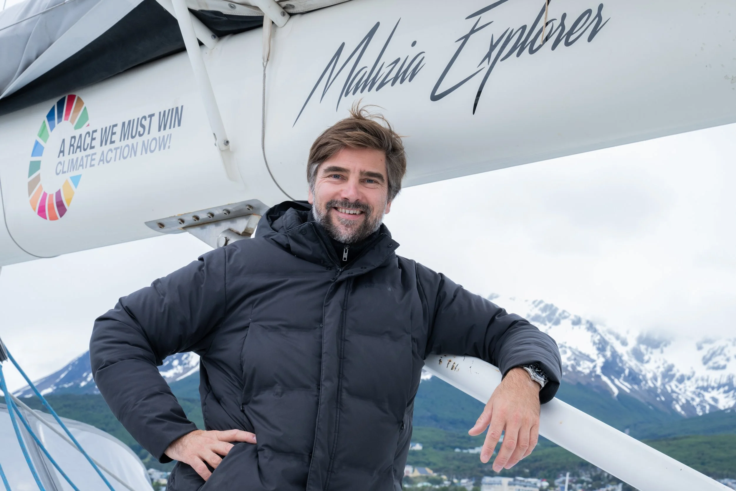 Boris Herrman, founder of Malizia Explorer. The sailboat has text that reads 'Malizia Explorer' and a slogan 'A RACE WE MUST WIN CLIMATE ACTION NOW!'. The man is smiling and leaning on the boat in Antarctica