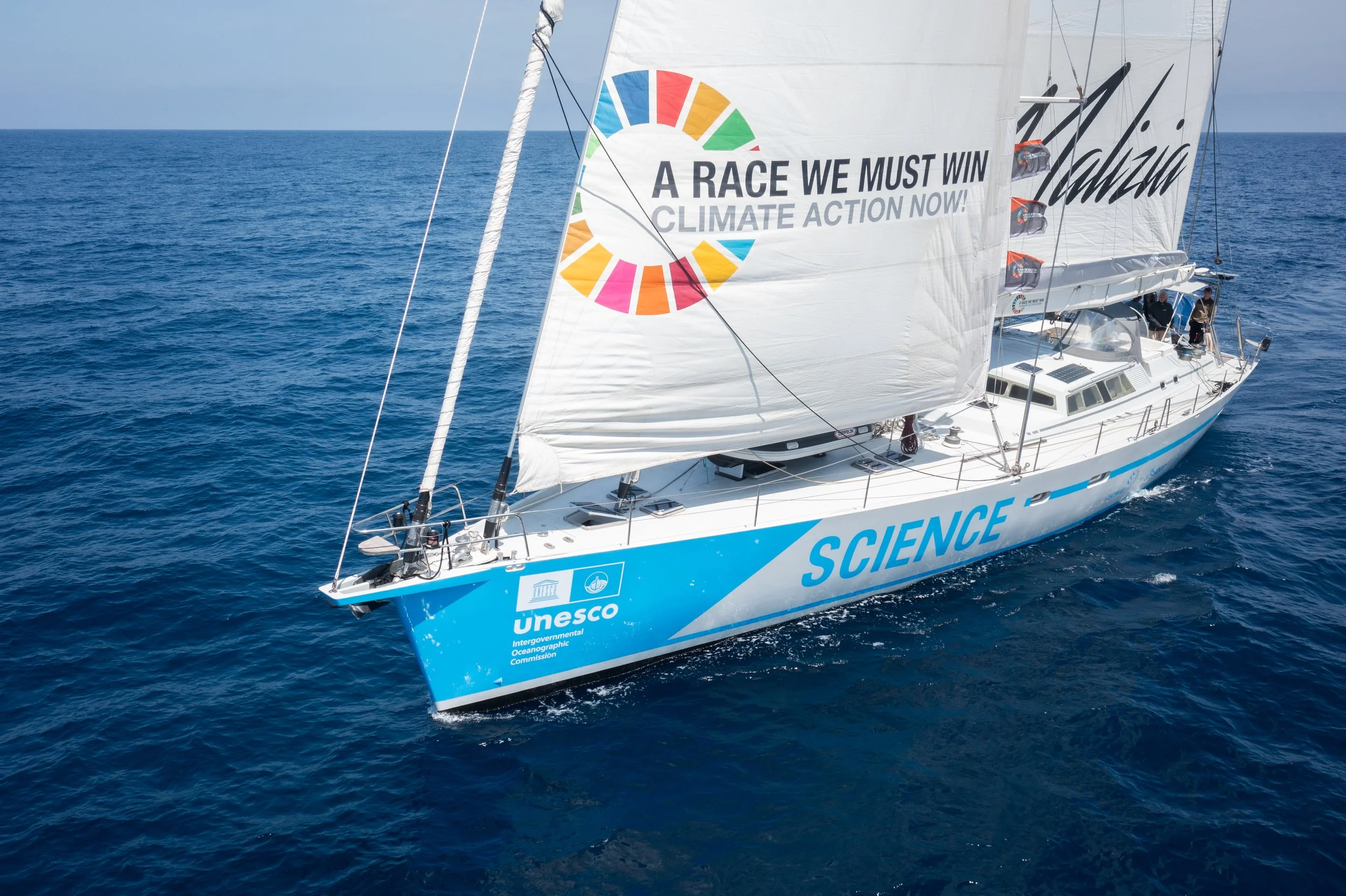 Malizia Explorer sailing yacht on the ocean with a large sail displaying a climate change awareness message. The yacht has the word 'SCIENCE' and the UNESCO logo on its side.