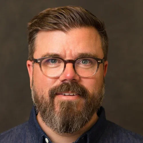 Close-up portrait of a man with glasses, a beard, and short hair, wearing a dark shirt, against a plain dark background.