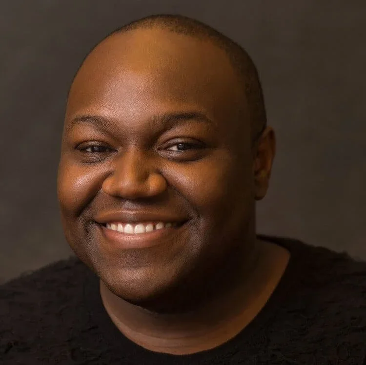 Close-up portrait of a smiling man with dark skin, wearing a black shirt against a neutral background.