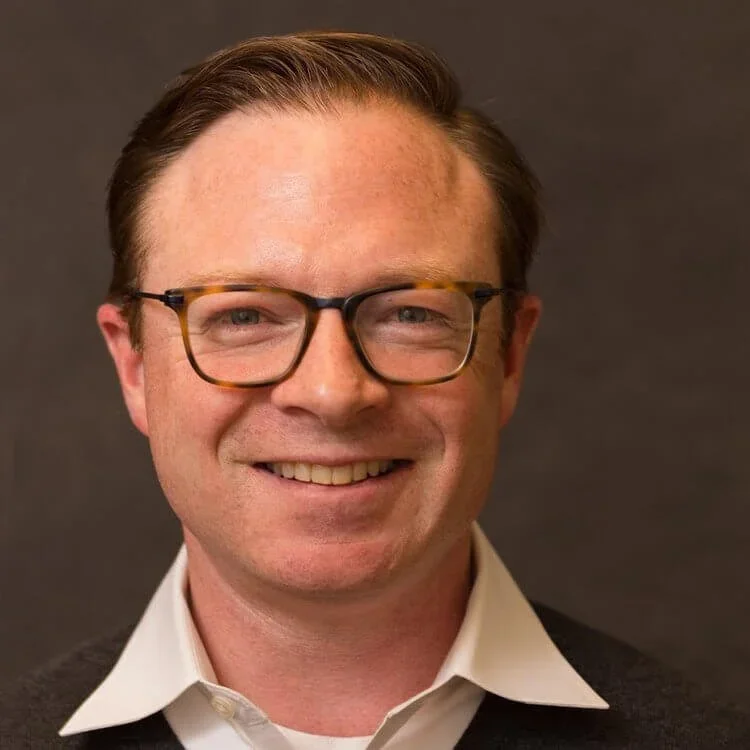 A smiling man wearing glasses, a white collared shirt, and a dark sweater against a dark background.