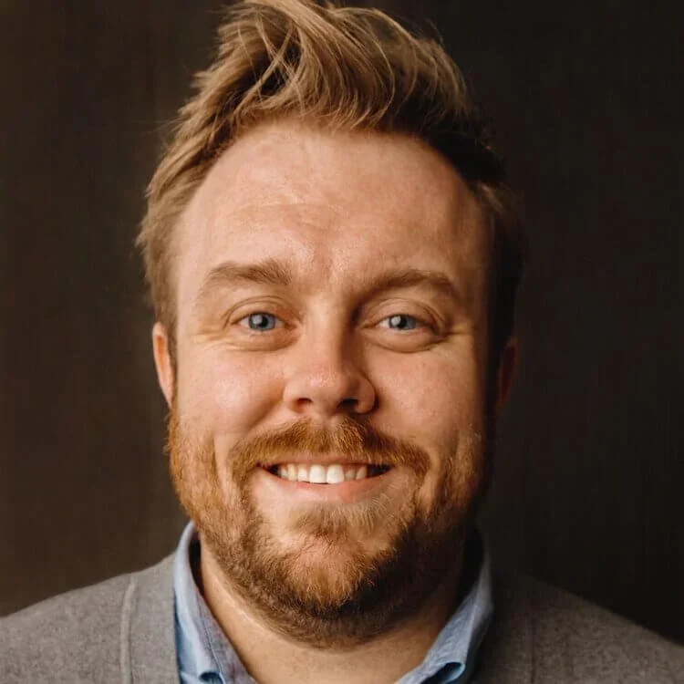 Portrait of a smiling man with light hair, beard, and blue eyes, wearing a gray jacket and blue shirt against a dark background.