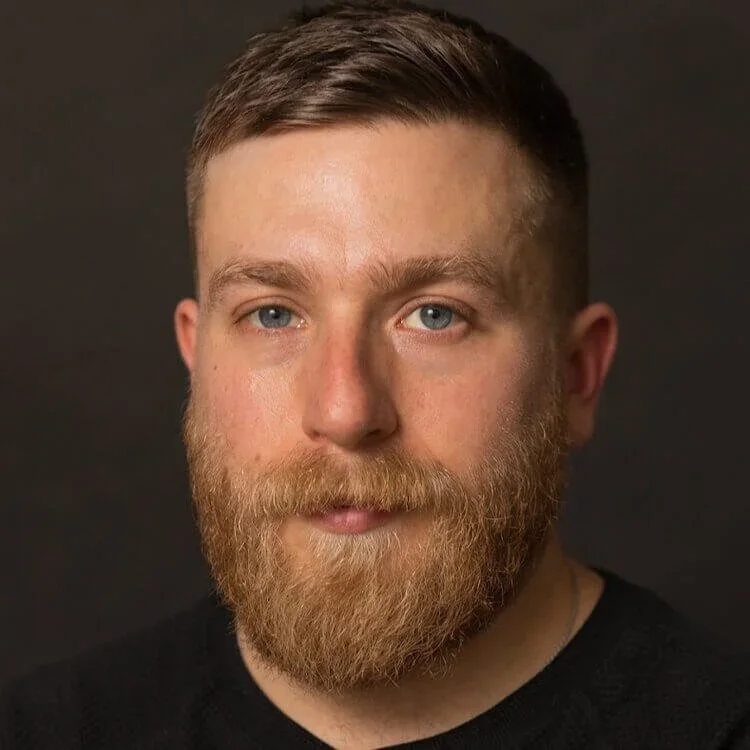 A close-up portrait of a man with short brown hair, a full red beard, and blue eyes, wearing a black shirt against a dark background.