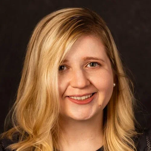 A close-up portrait of a young woman with blonde hair, smiling, wearing pearl earrings against a dark background.