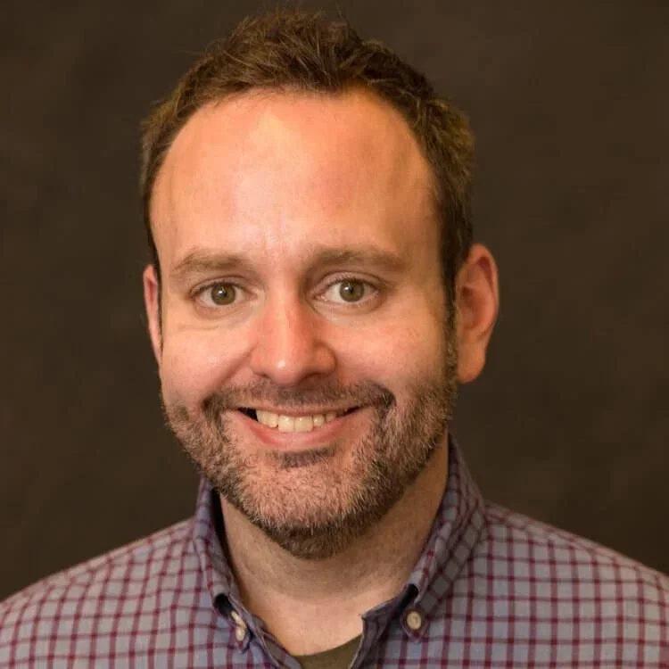 A close-up of a middle-aged man with light brown hair and a beard, smiling and wearing a checkered shirt, against a dark background.