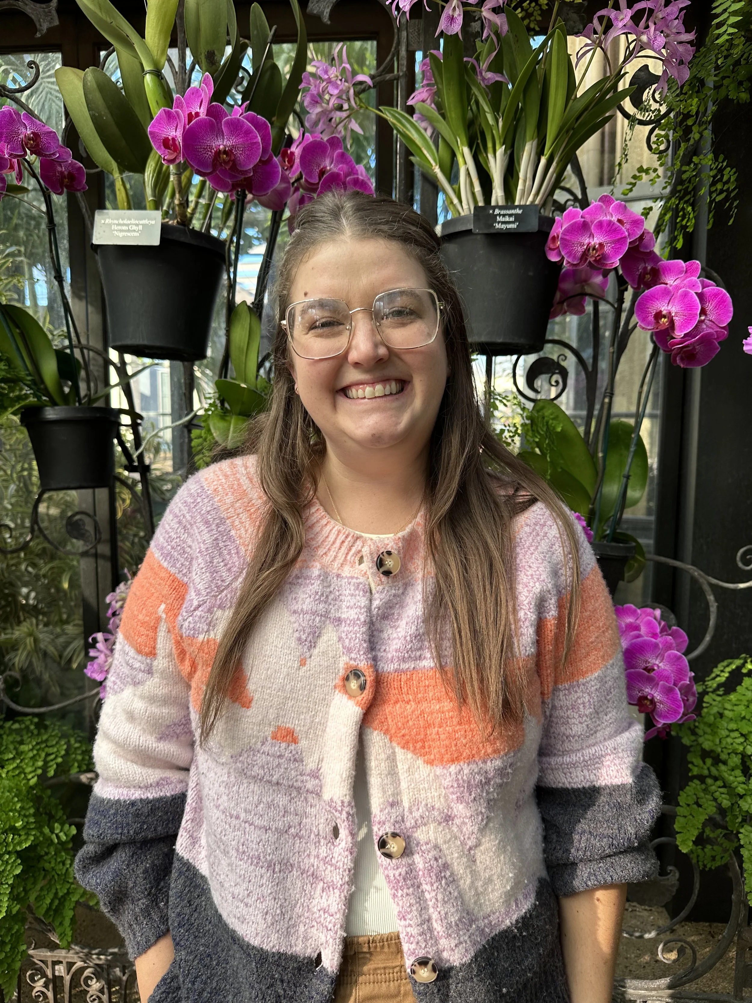 A woman with long brown hair and glasses smiling in front of a display of pink orchids in a greenhouse or botanical garden.