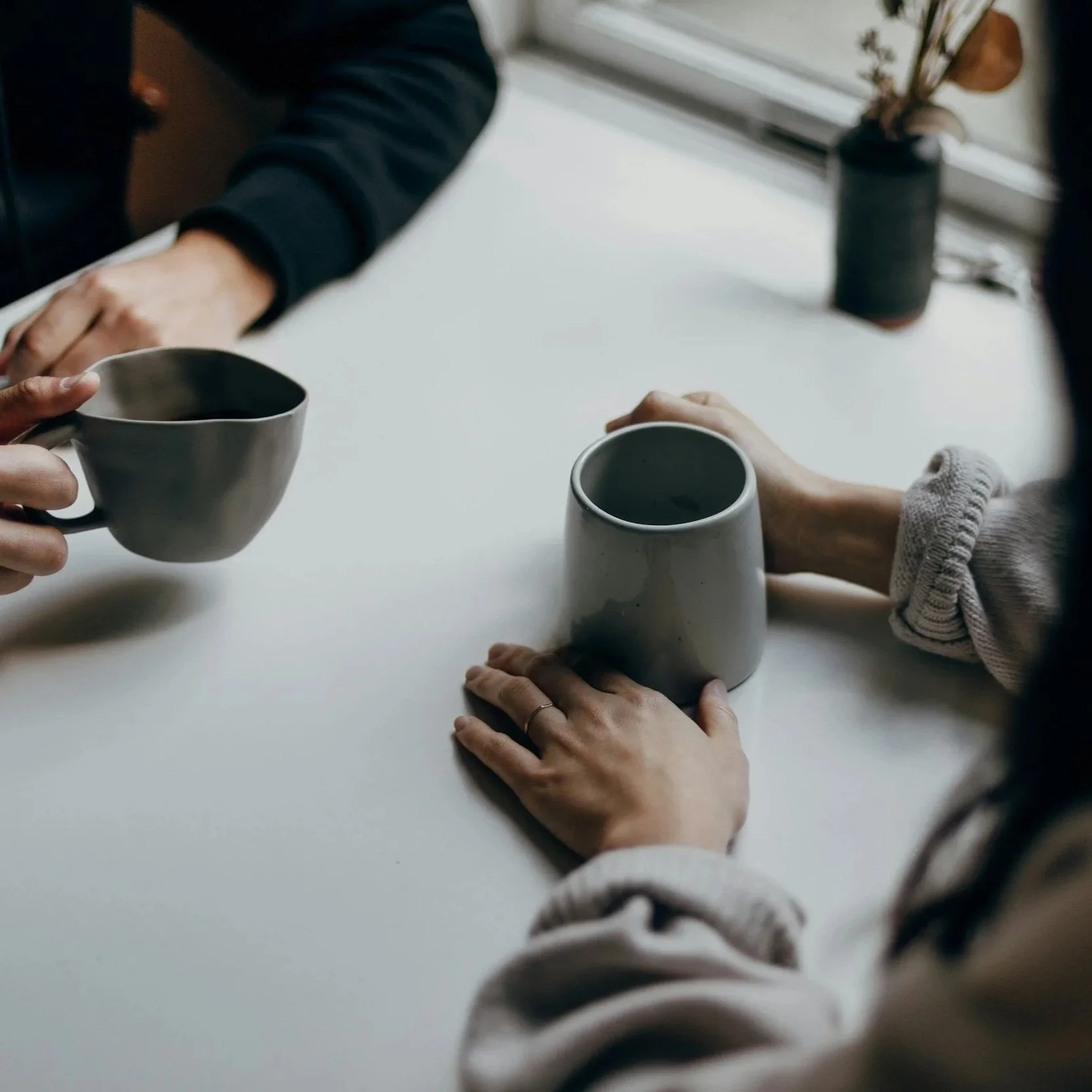Two people exchanging coffee mugs at a white table, with a potted plant in the background.