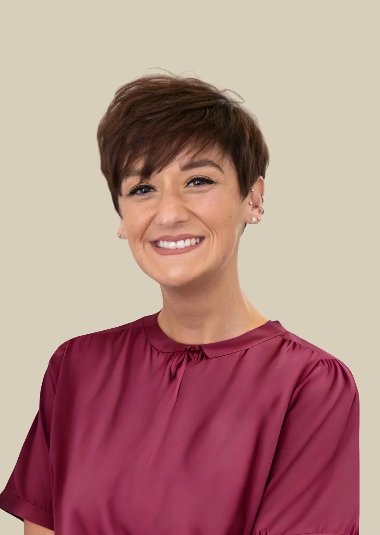 A woman with short, dark brown hair styled in a pixie cut, smiling and wearing a shiny, burgundy satin blouse against a plain, light-colored background.