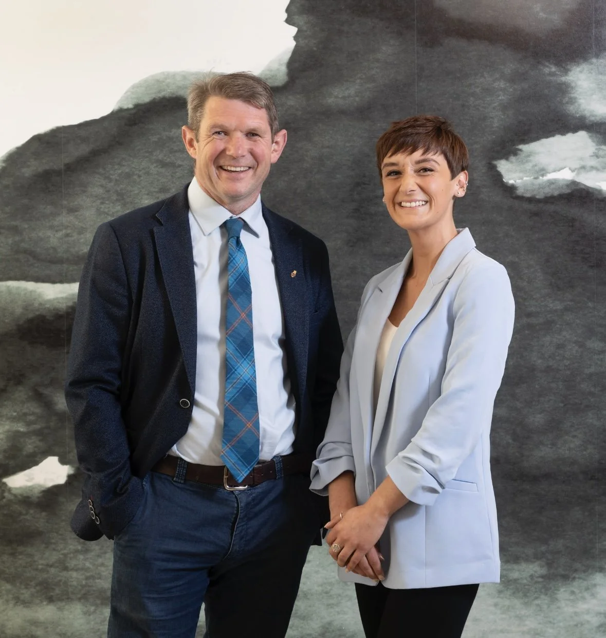 A man and a woman in business attire standing in front of an abstract black and white mural, smiling at the camera.