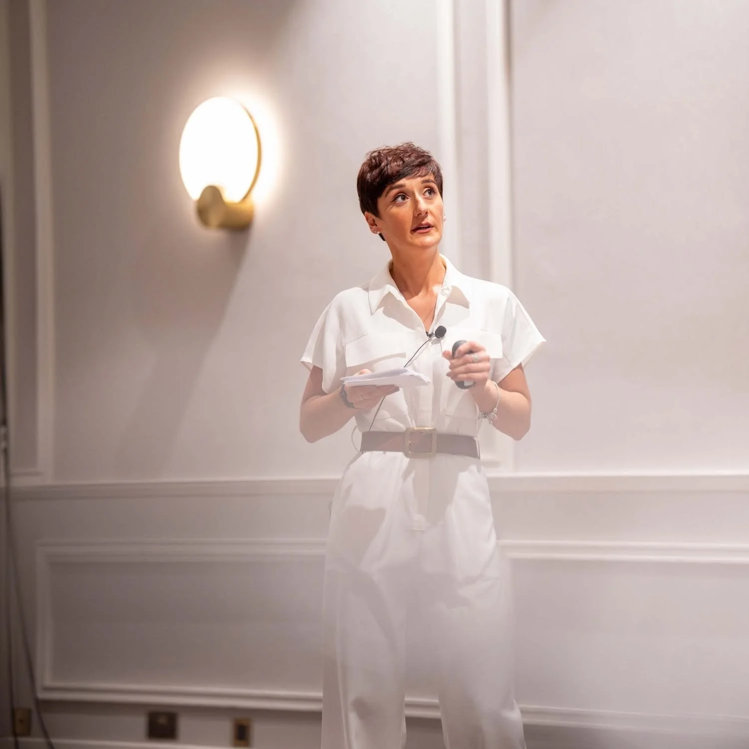 A woman with short dark hair, wearing an all-white outfit, standing in front of a white wall with a gold wall sconce. She appears to be speaking or giving a presentation, holding notes and a microphone.